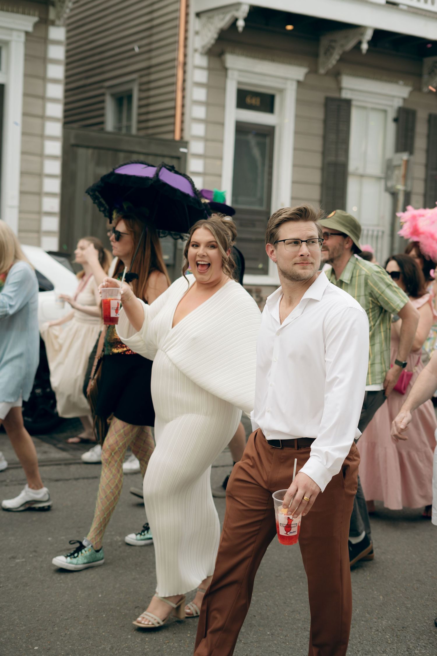 A couple strolls together at an outdoor event, with the woman wearing a white dress and man in brown pants and white shirt.