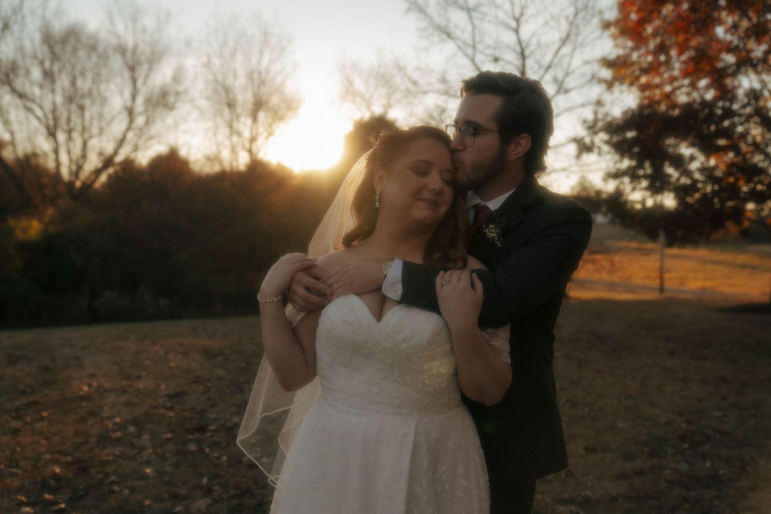 A romantic sunset wedding photo of a couple embracing in a field with golden light filtering through autumn trees.