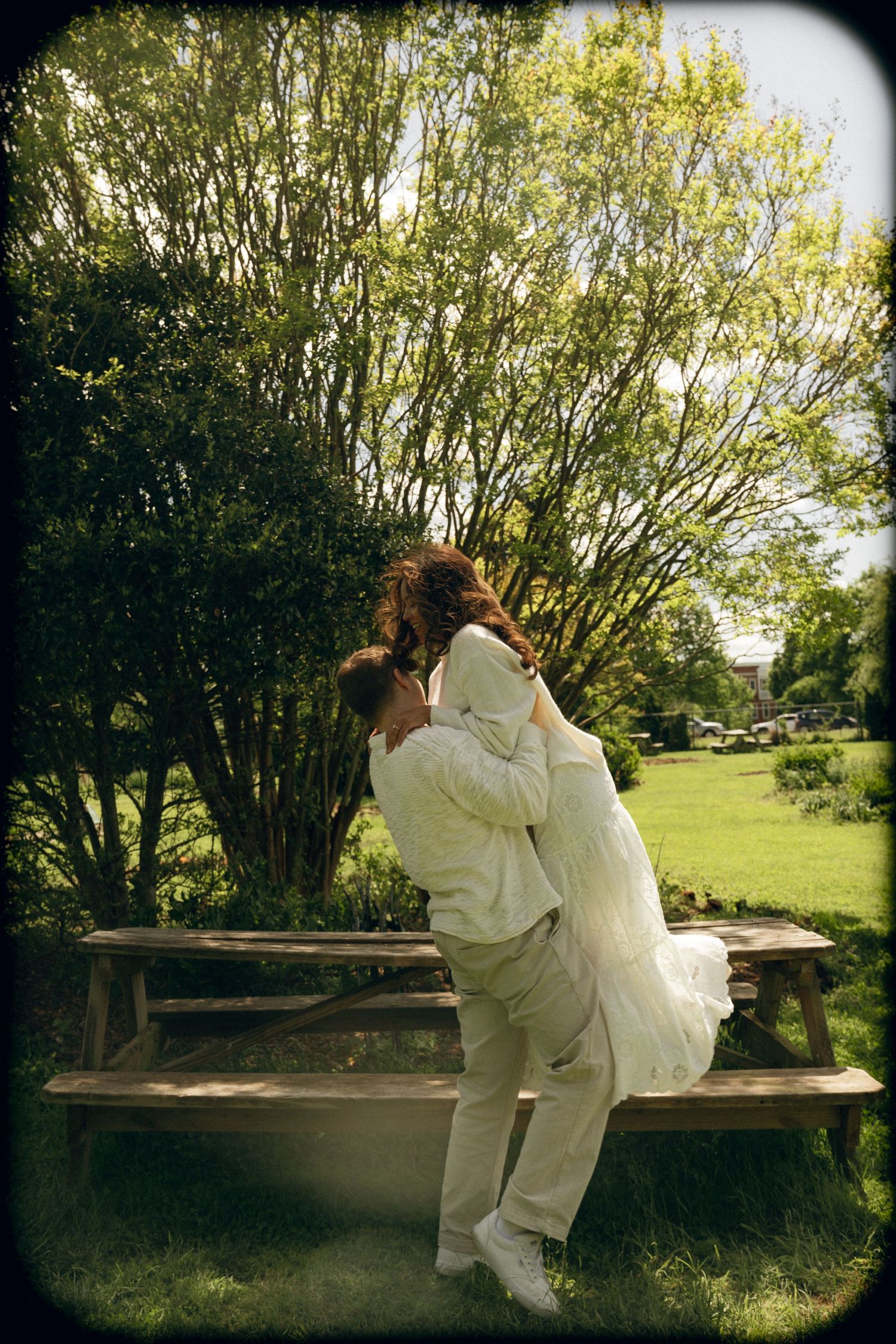 Bride and groom embracing near a picnic table in a lush green garden on their wedding day.