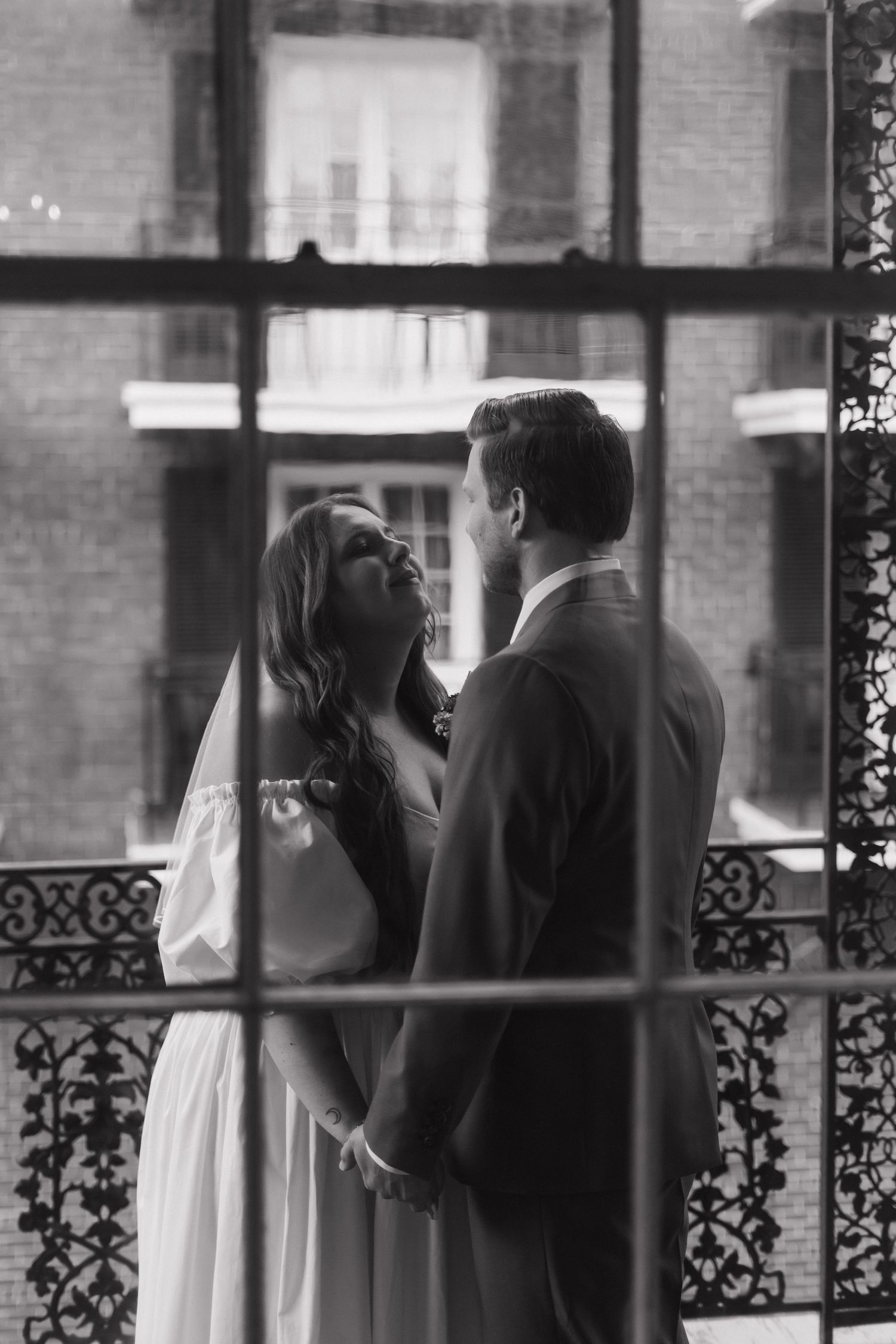 A romantic black and white photograph of a couple on a balcony with ornate iron railings, captured through window panes.