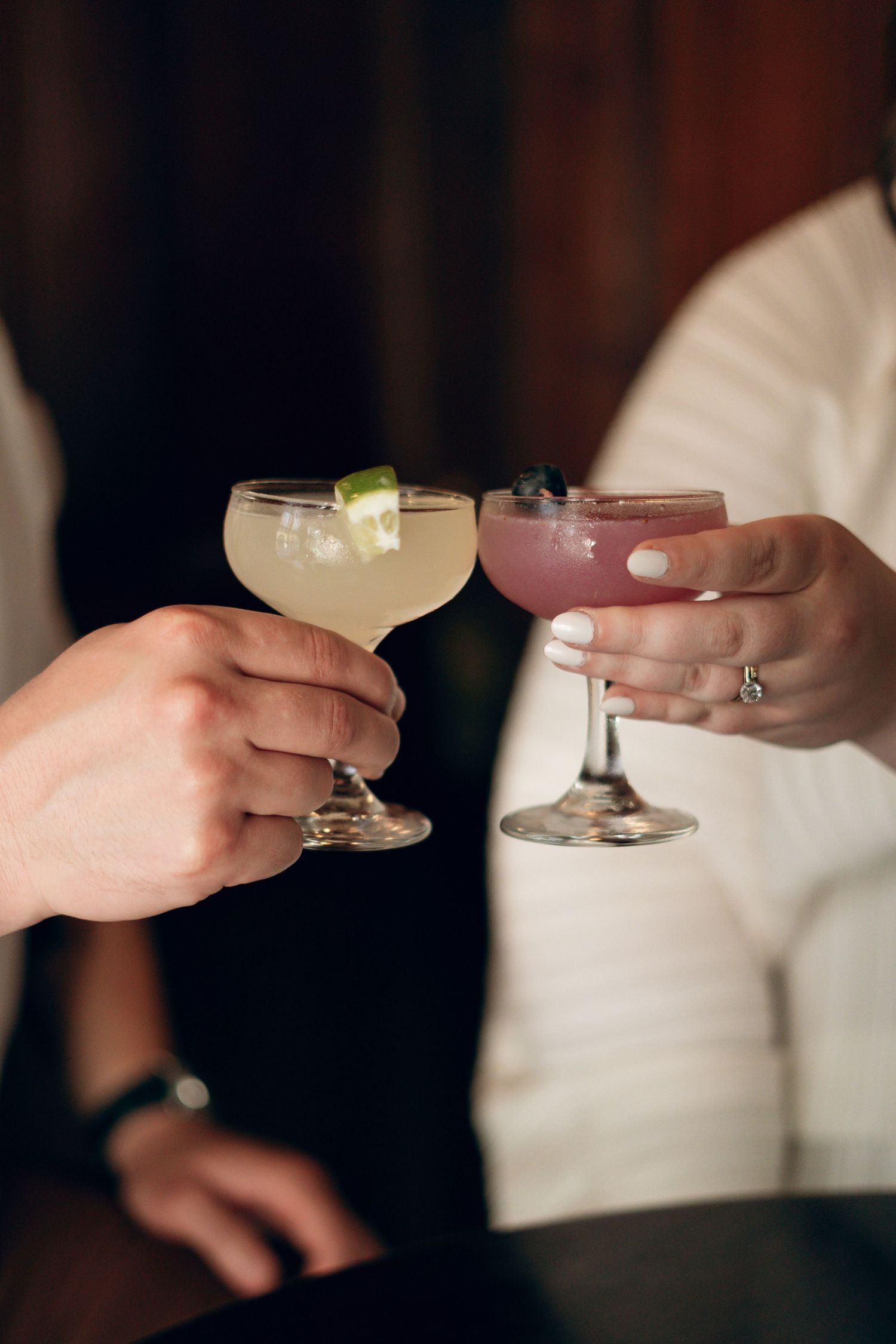 Two people clink cocktail glasses in an intimate toast, with one clear and one pink drink garnished with lime.