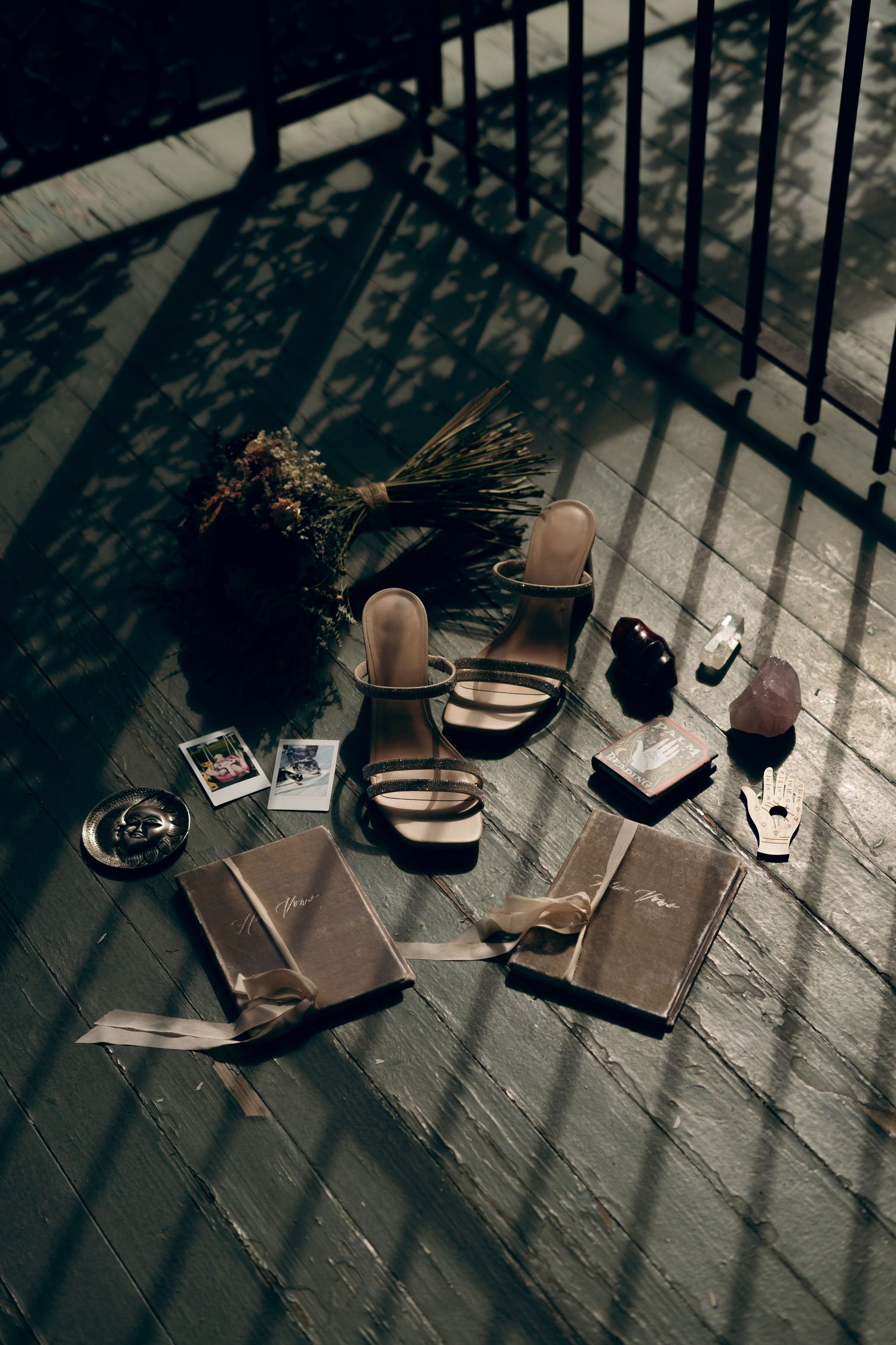 A moody still life of sandals, books, and a dried flower arrangement on a wooden deck with dramatic shadows.