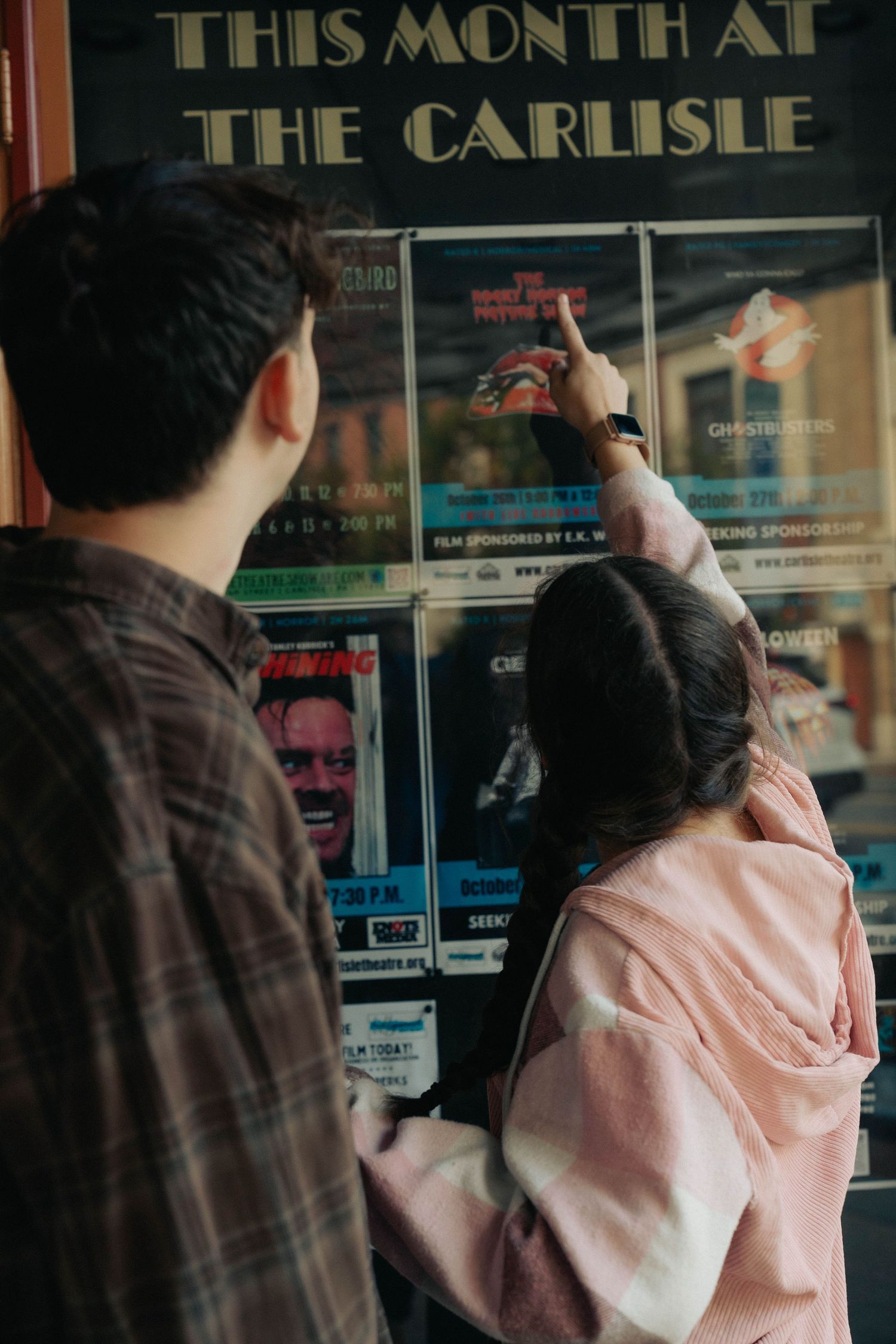 Two people look at event posters on a wall under a sign reading 'THIS MONTH AT THE CARLISLE'.