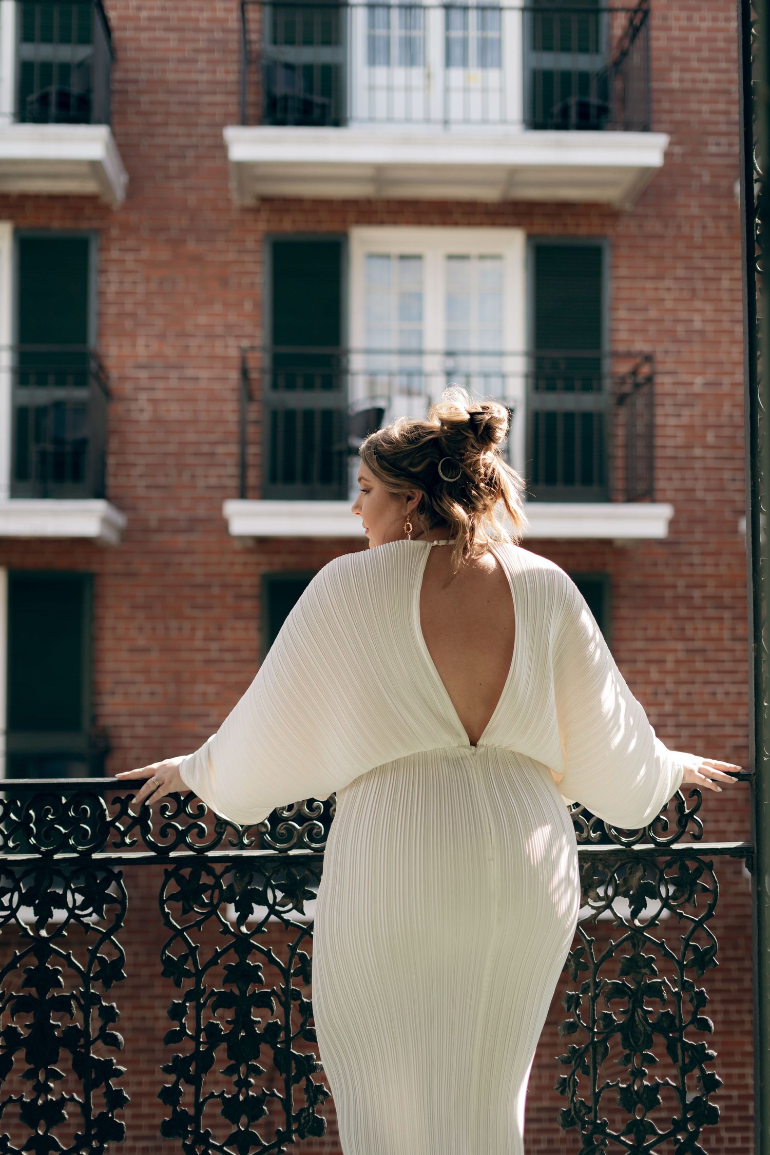 A person in a white backless dress stands on a balcony overlooking a brick apartment building with ornate railings.