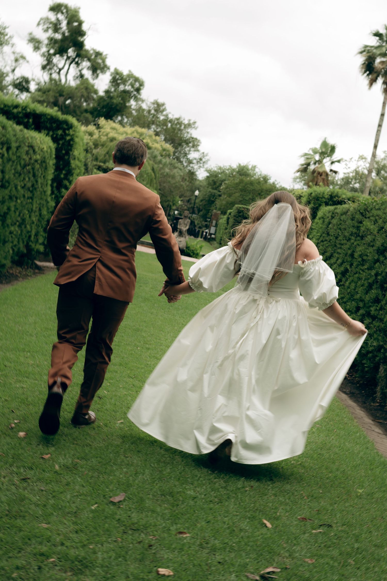 A couple runs joyfully through a garden pathway, with the bride in a flowing white gown and groom in brown attire.