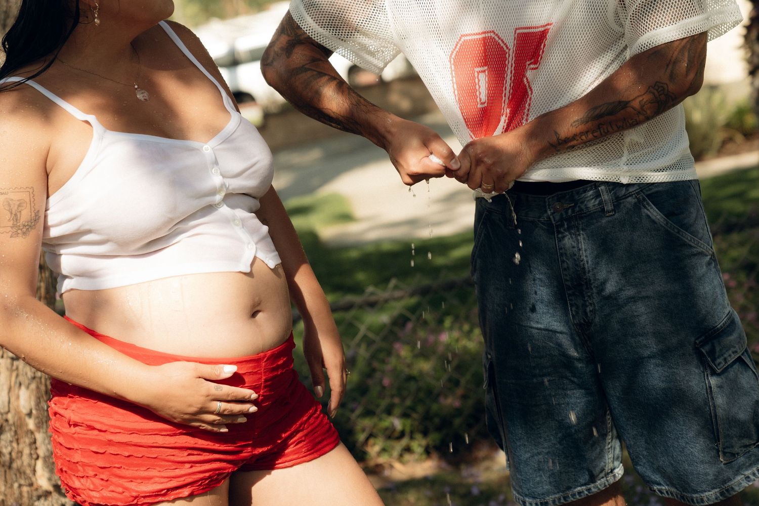 A pregnant woman in white shirt and pink pants stands outdoors while someone touches her baby bump.