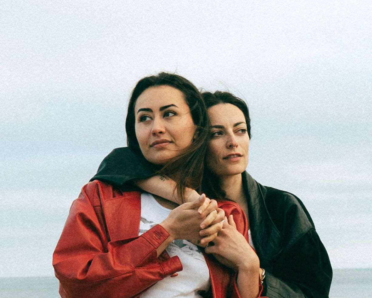 Two people embrace outdoors, one wearing a red jacket and one in black, against a cloudy sky background.