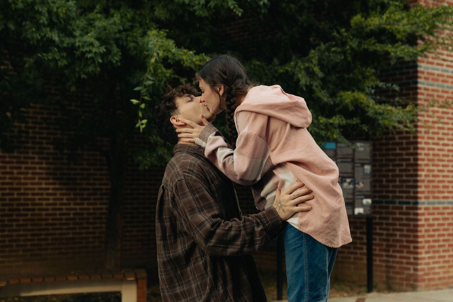 Two people share an intimate moment in the rain outside a brick building while one wears a pink hoodie and another a brown jacket.