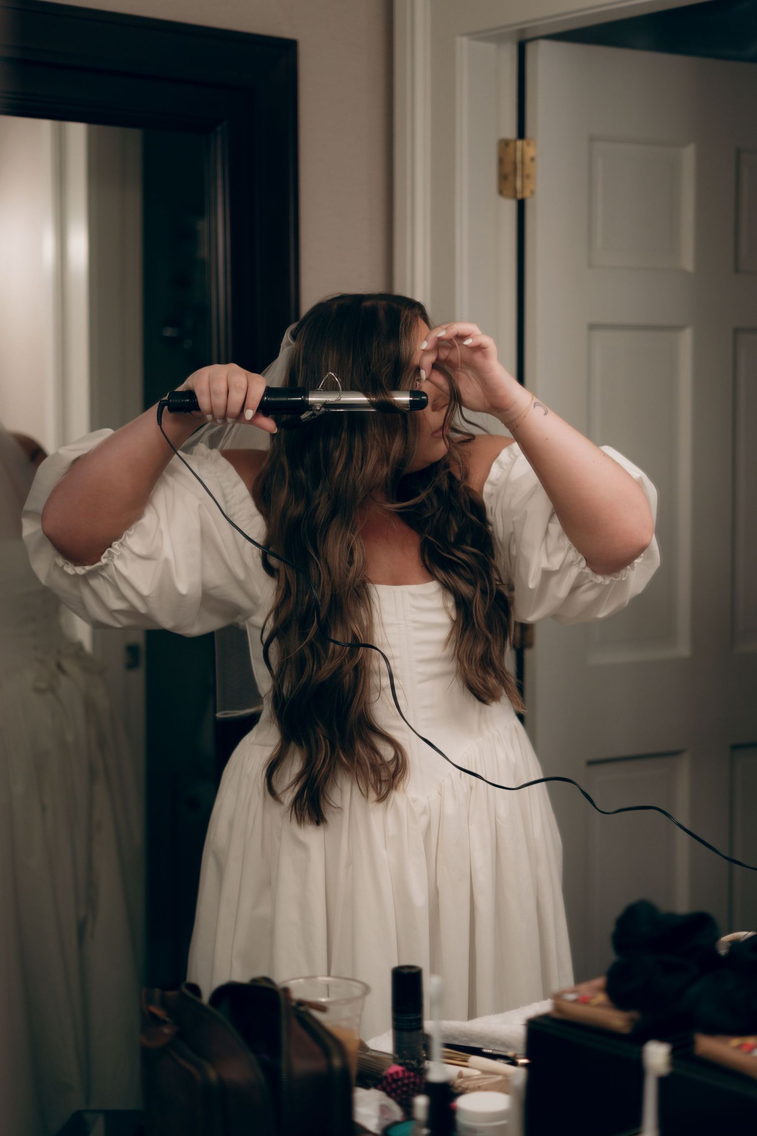 A woman in a white dress styles her long dark hair using a curling iron while standing in a bathroom.