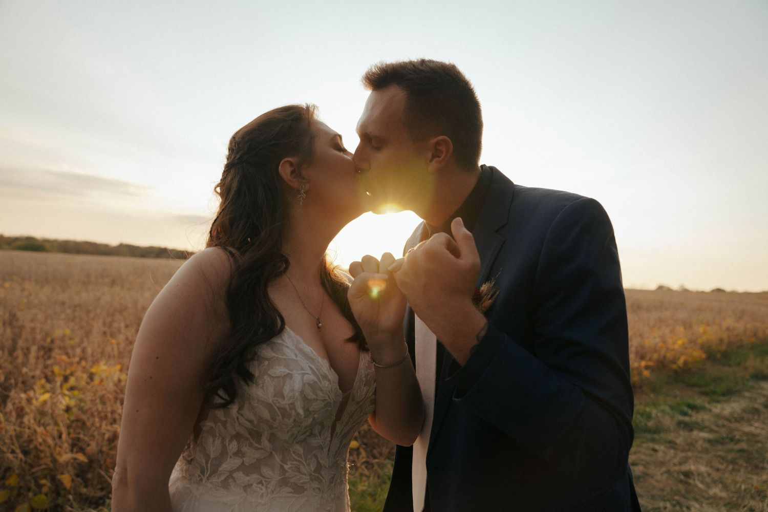 A tender kiss between newlyweds silhouetted against a glowing sunset in a field.