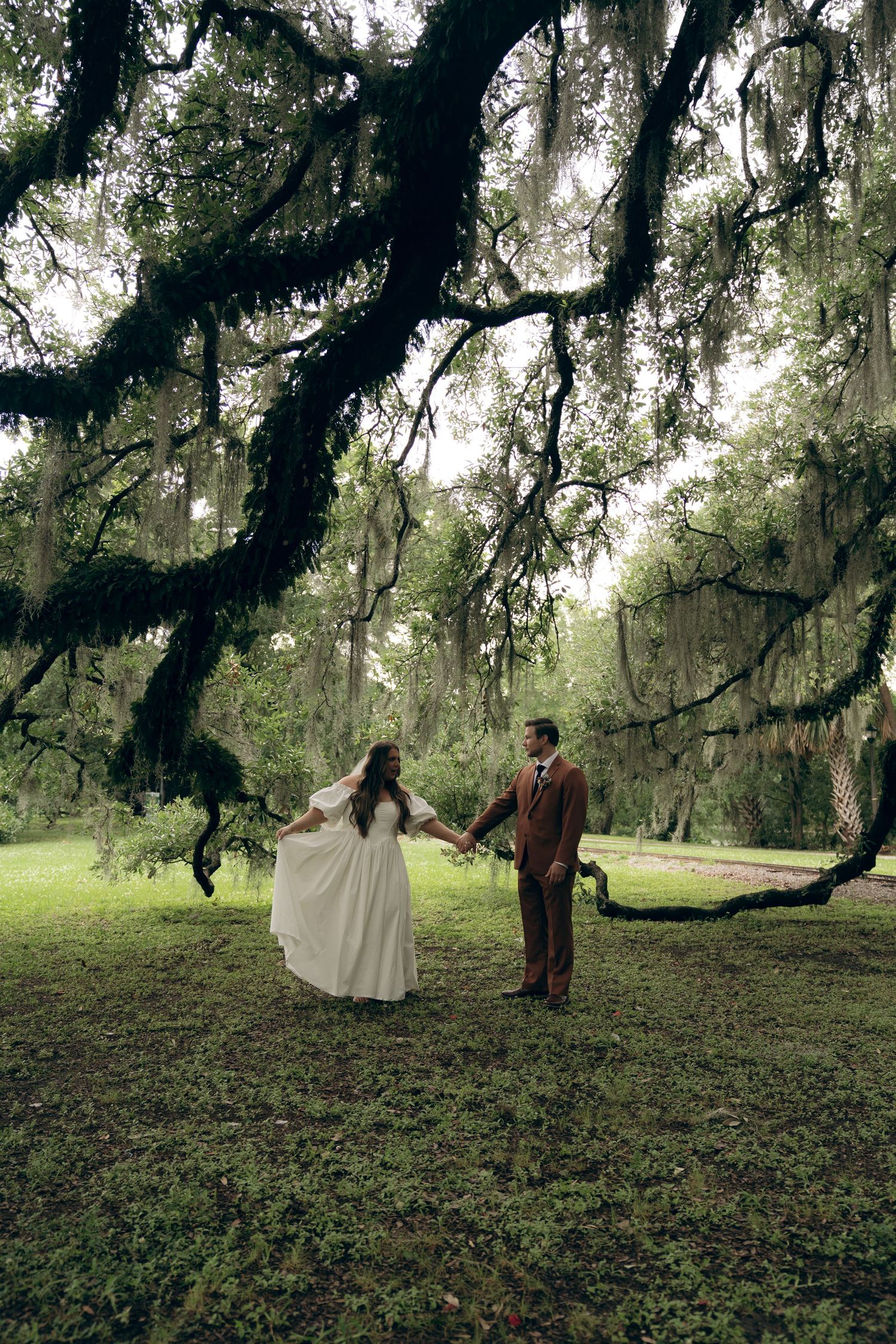 A couple stands beneath majestic Spanish moss-draped oak trees in a dreamy Southern garden setting.