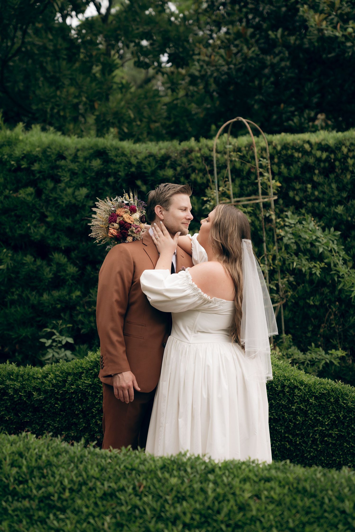 A wedding couple stands in an outdoor garden setting surrounded by lush greenery and hedges.