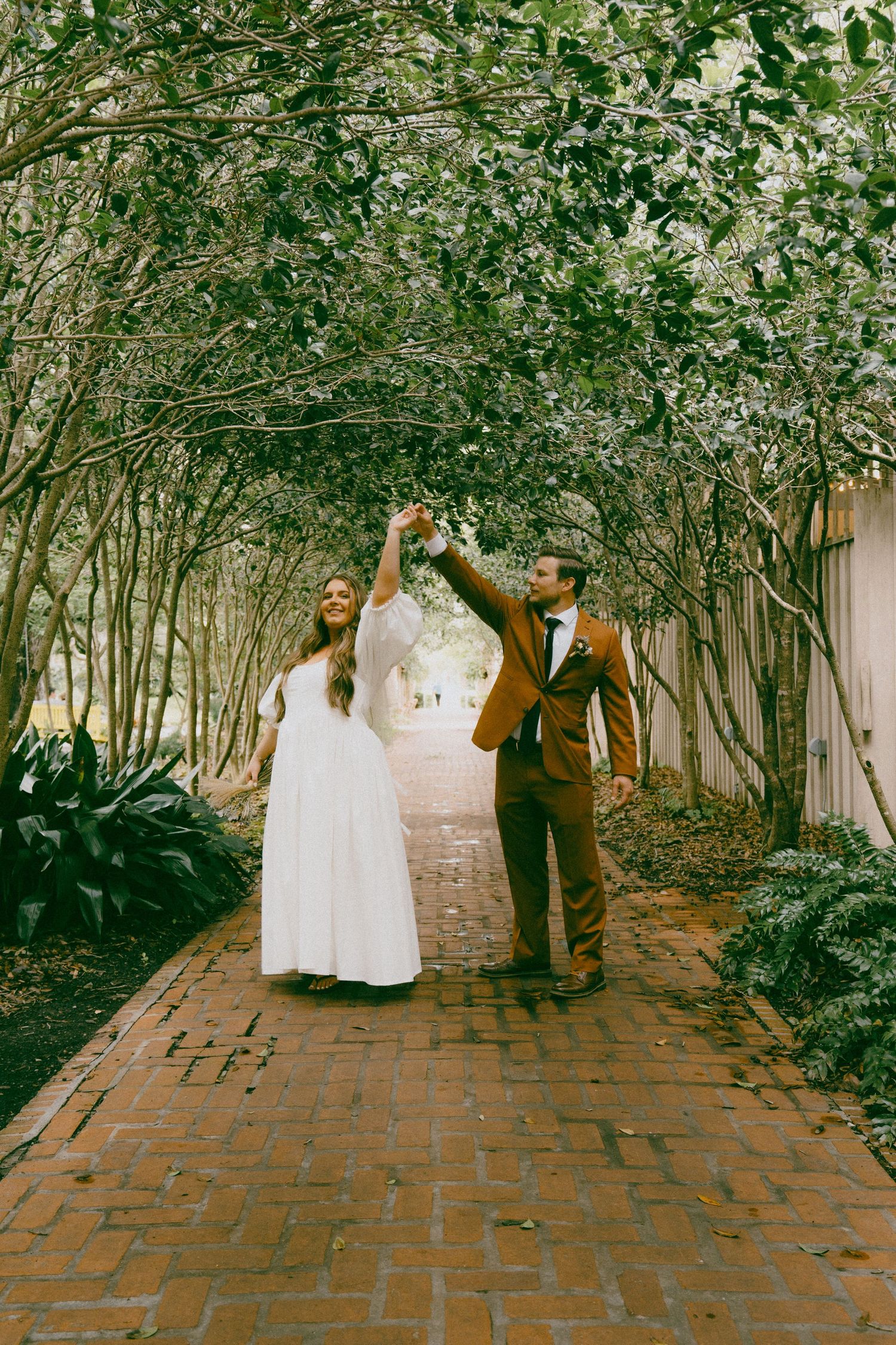 Four individuals in formal attire walking under a decorative archway of green leaves and branches on a brick path.