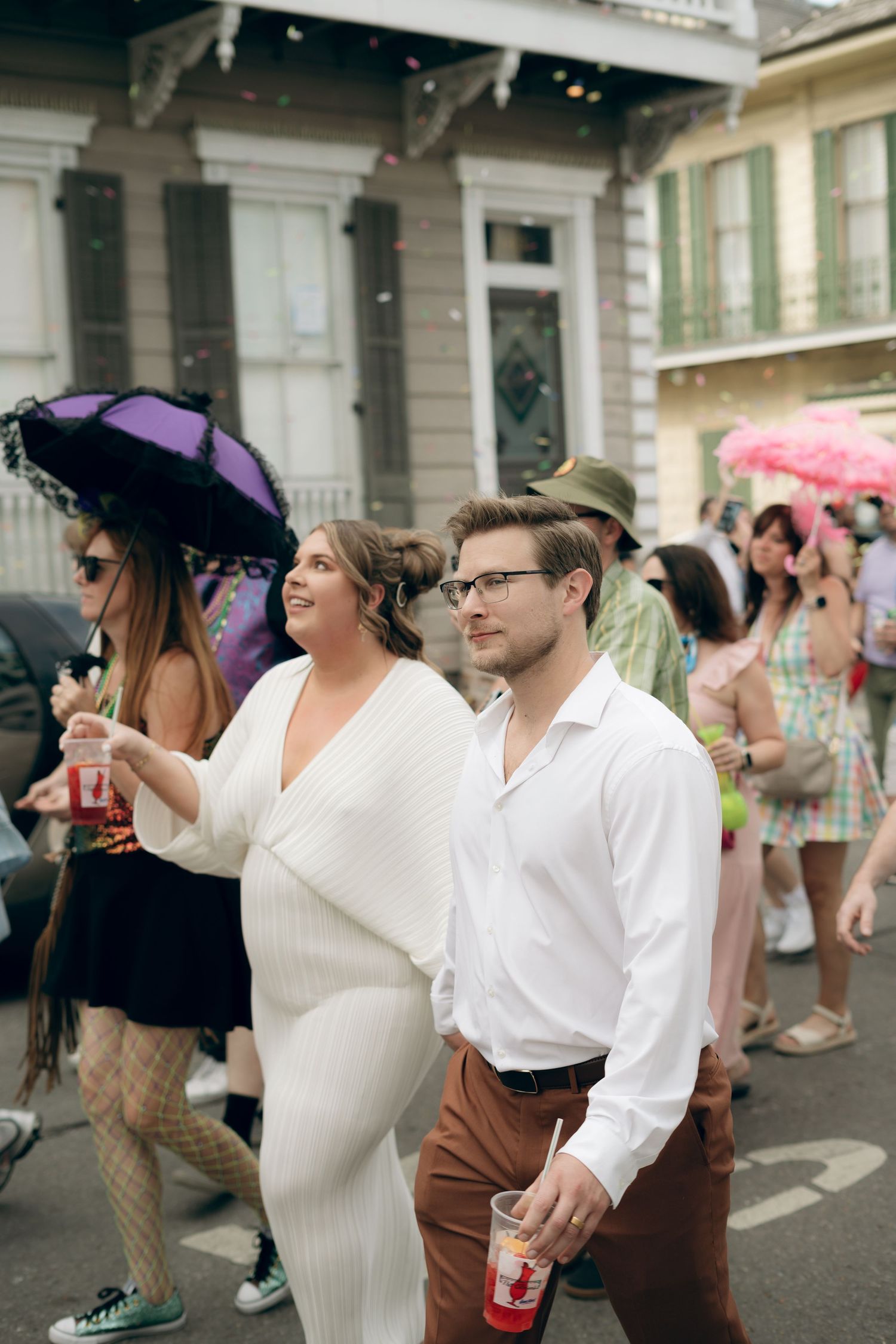 A festive scene with celebrants in white attire enjoying drinks and parasols at a New Orleans street party.