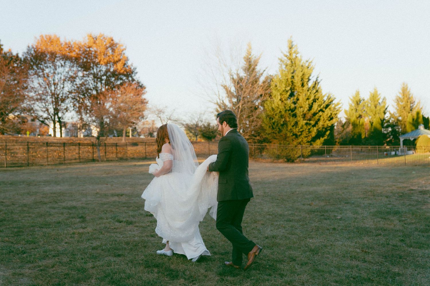 Newlyweds walk together across lawn during autumn sunset.