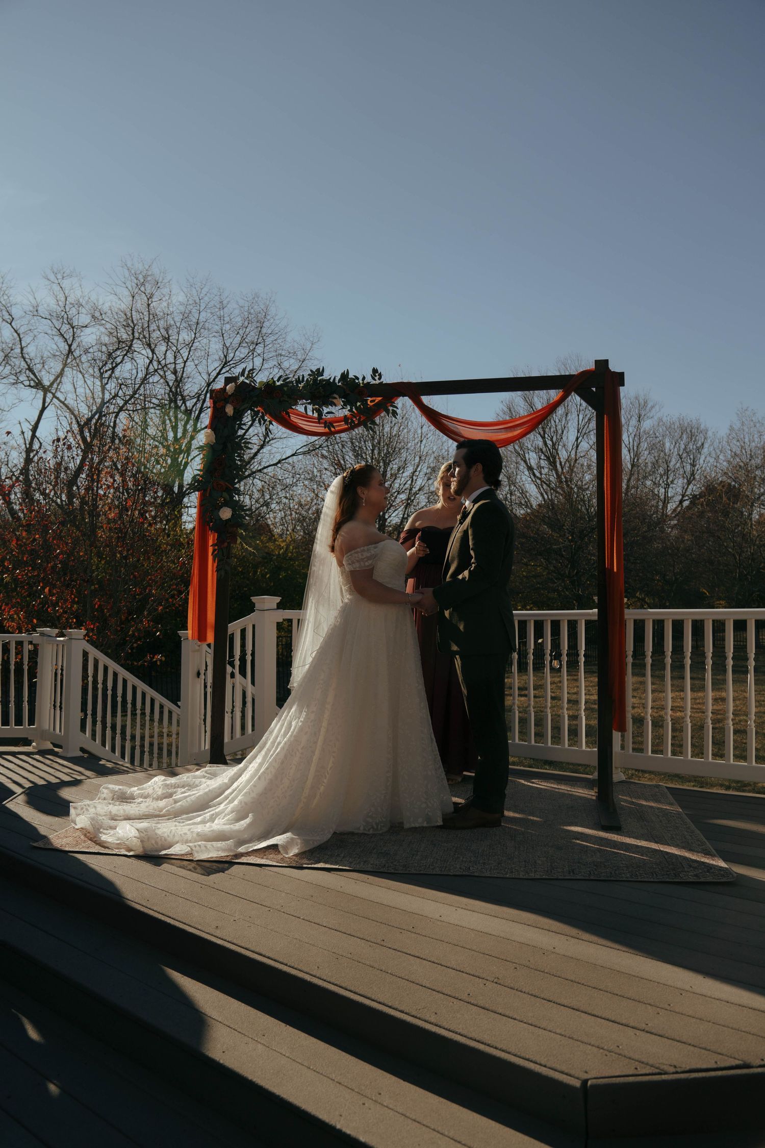 A wedding ceremony taking place on a wooden deck with orange draped arch and autumn foliage in background.