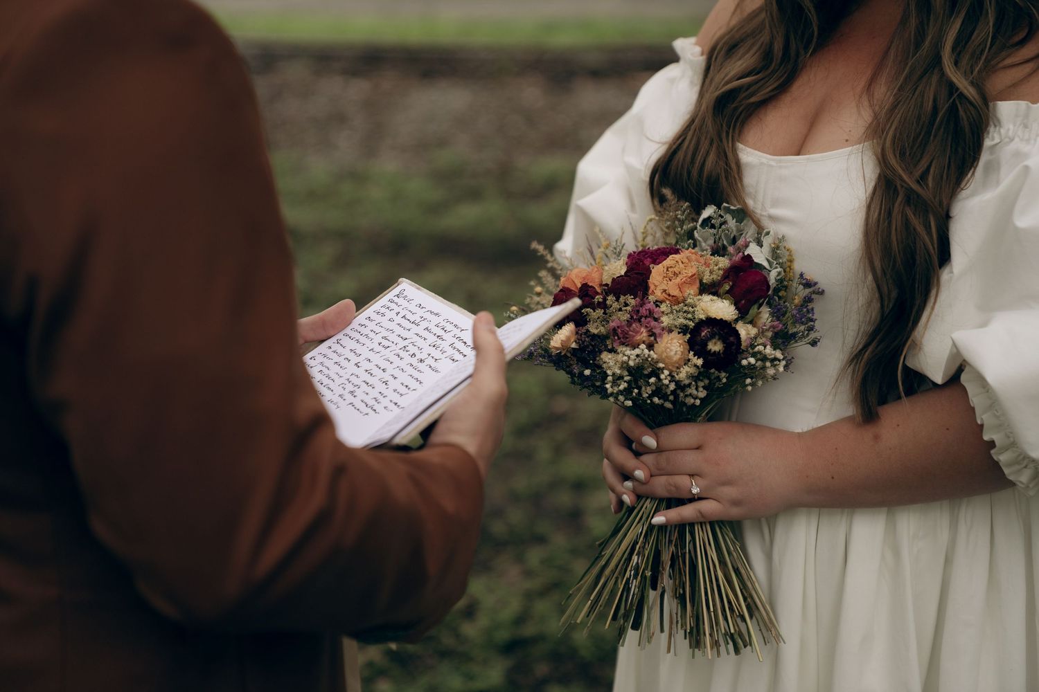Close-up of bride holding bouquet while groom reads handwritten vows during outdoor wedding ceremony.