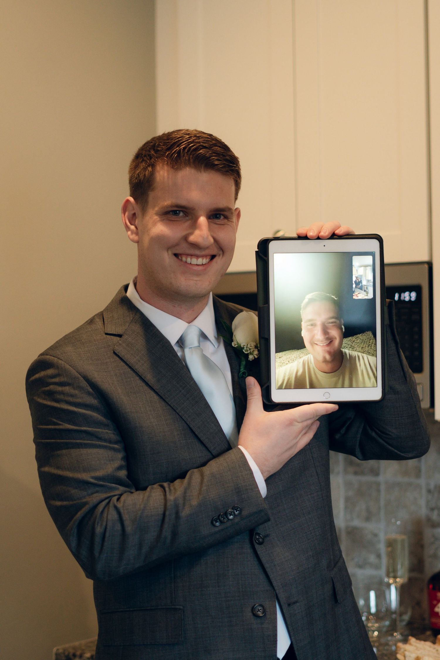 A groom in a gray suit shows a video call on a tablet during wedding preparations.