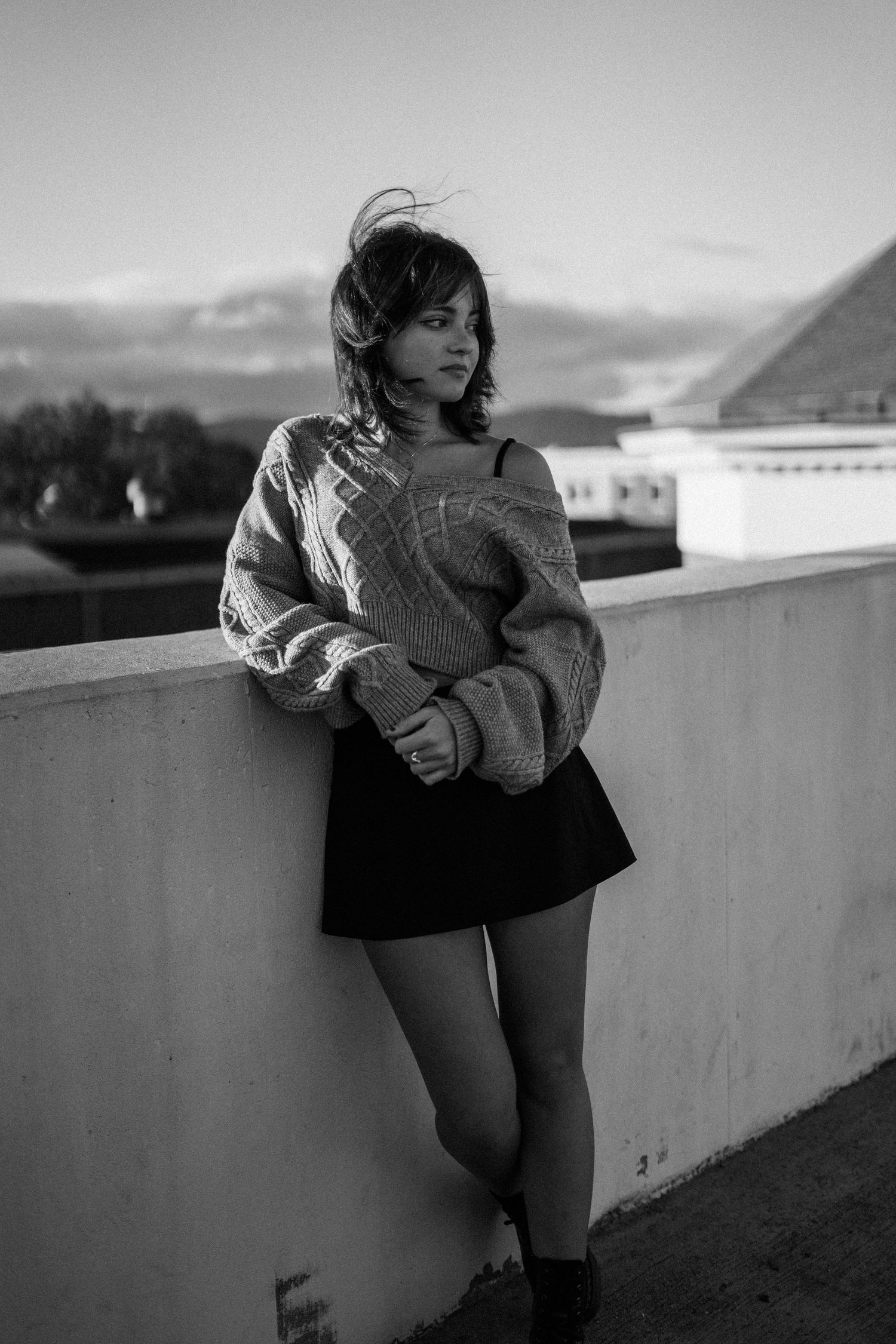 Woman in cozy sweater and skirt poses against wall with mountains in background, black and white photography.