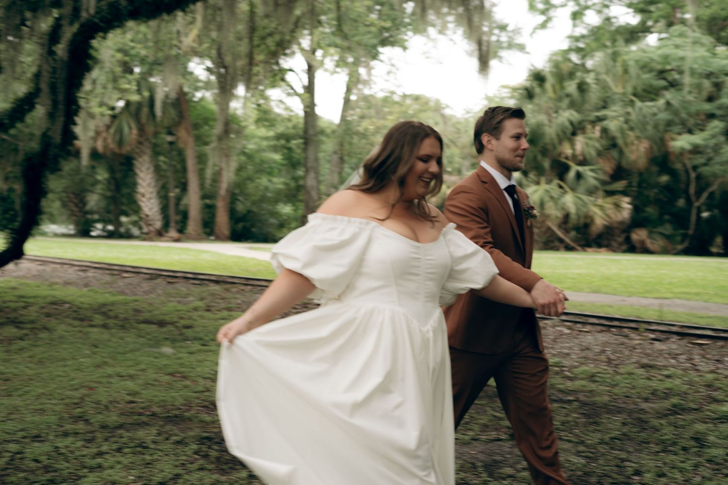 A couple dances joyfully in a garden setting, with the person in white wearing a flowing off-shoulder dress.