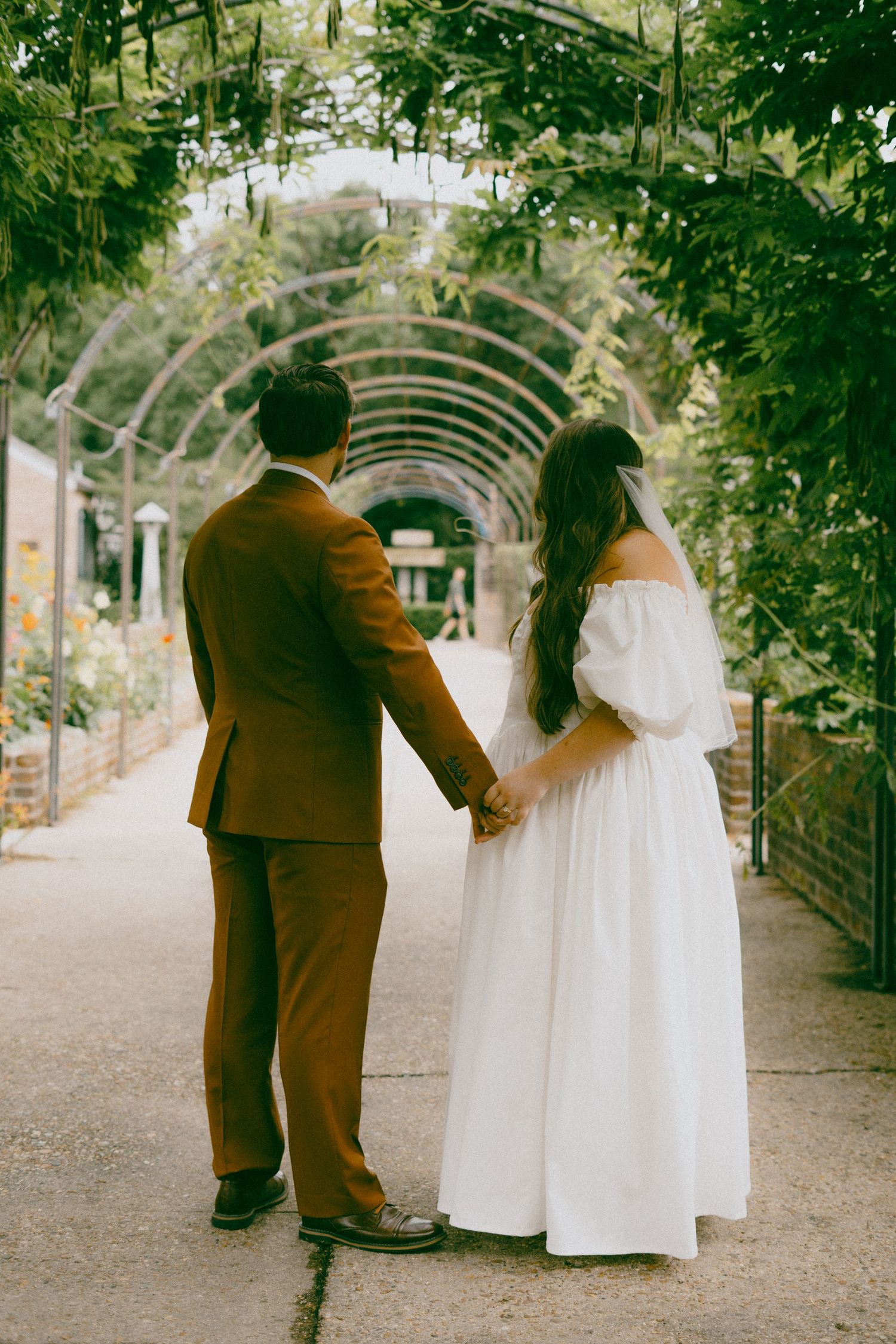 A couple in wedding attire holds hands while walking through a romantic garden archway covered in greenery.