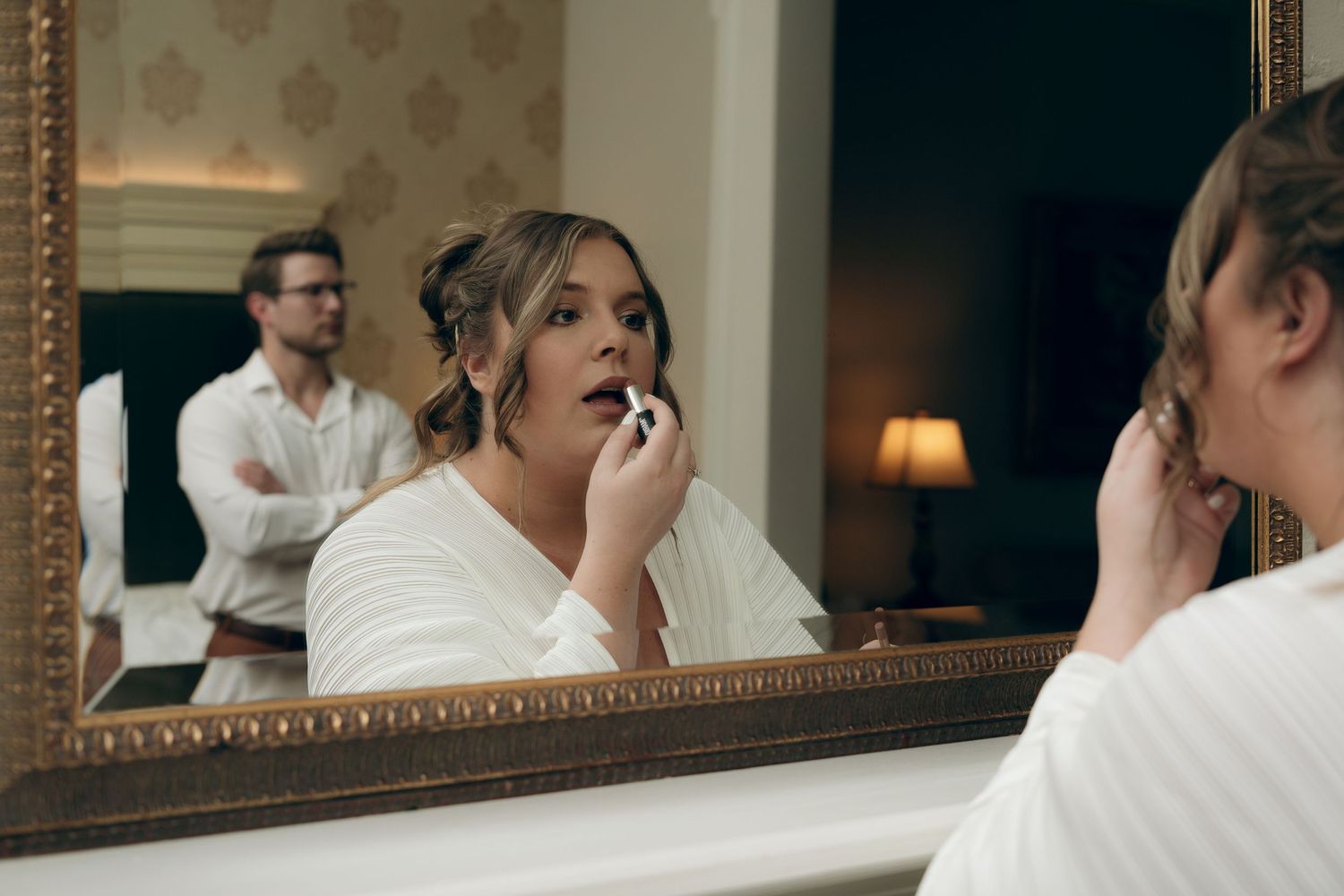 A person in a white robe applies makeup while looking into a framed mirror with another figure in background.