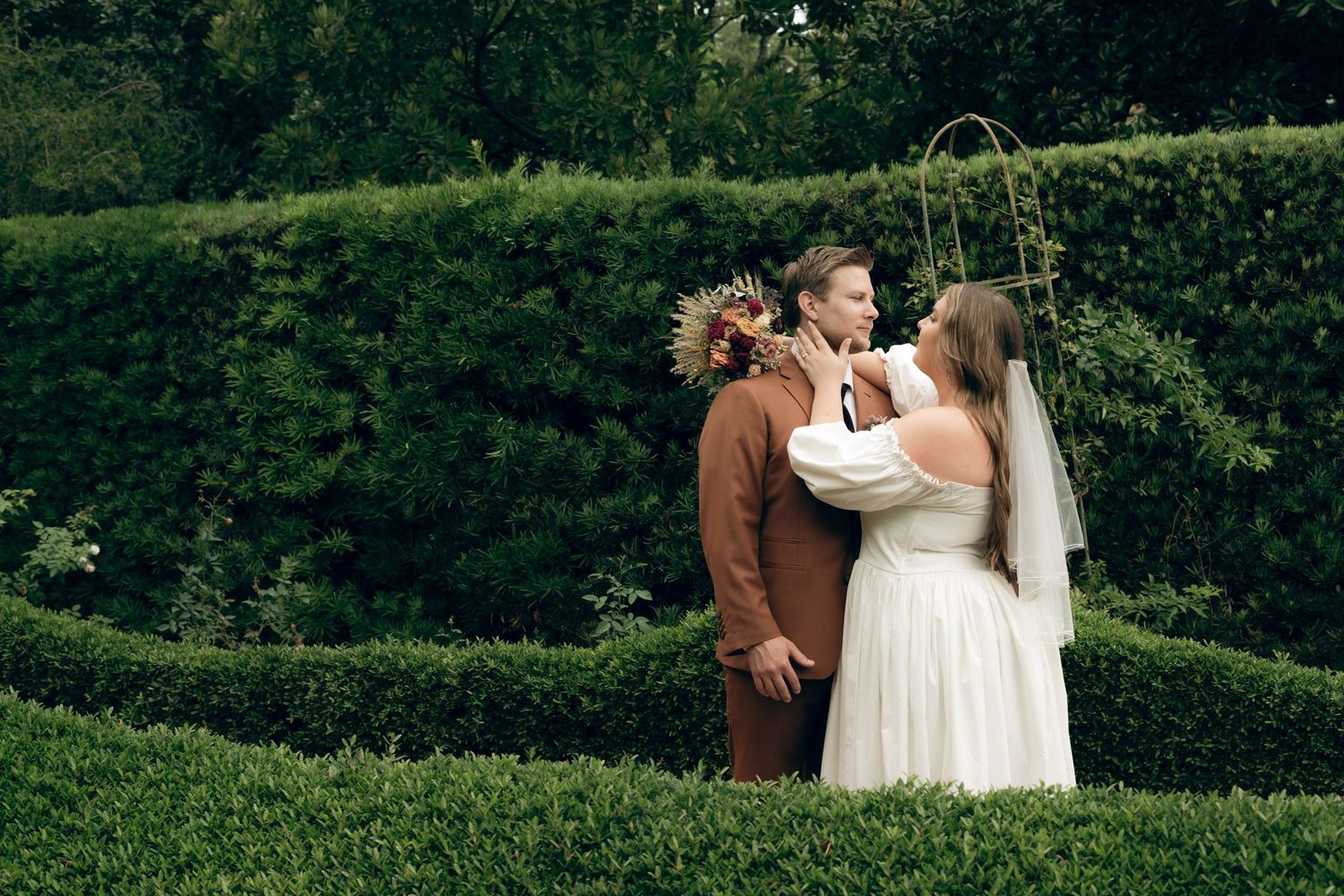 A romantic couple embrace in a garden maze with lush green hedges as their backdrop on their wedding day.