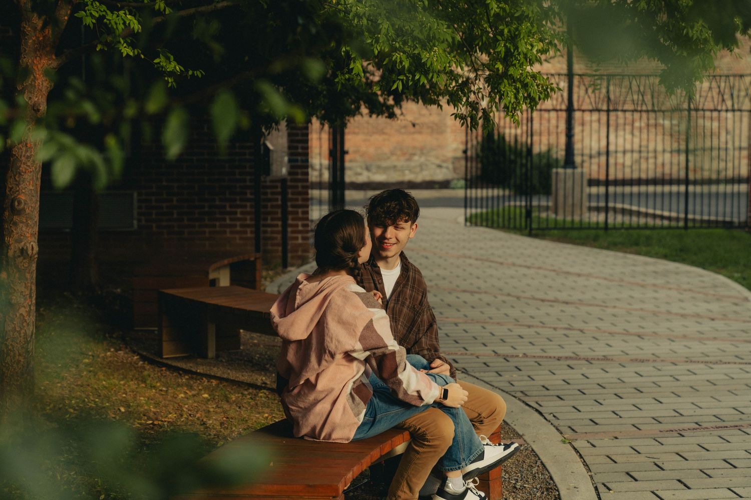 Two people sit close together on a park bench along a curved brick pathway on a peaceful evening.