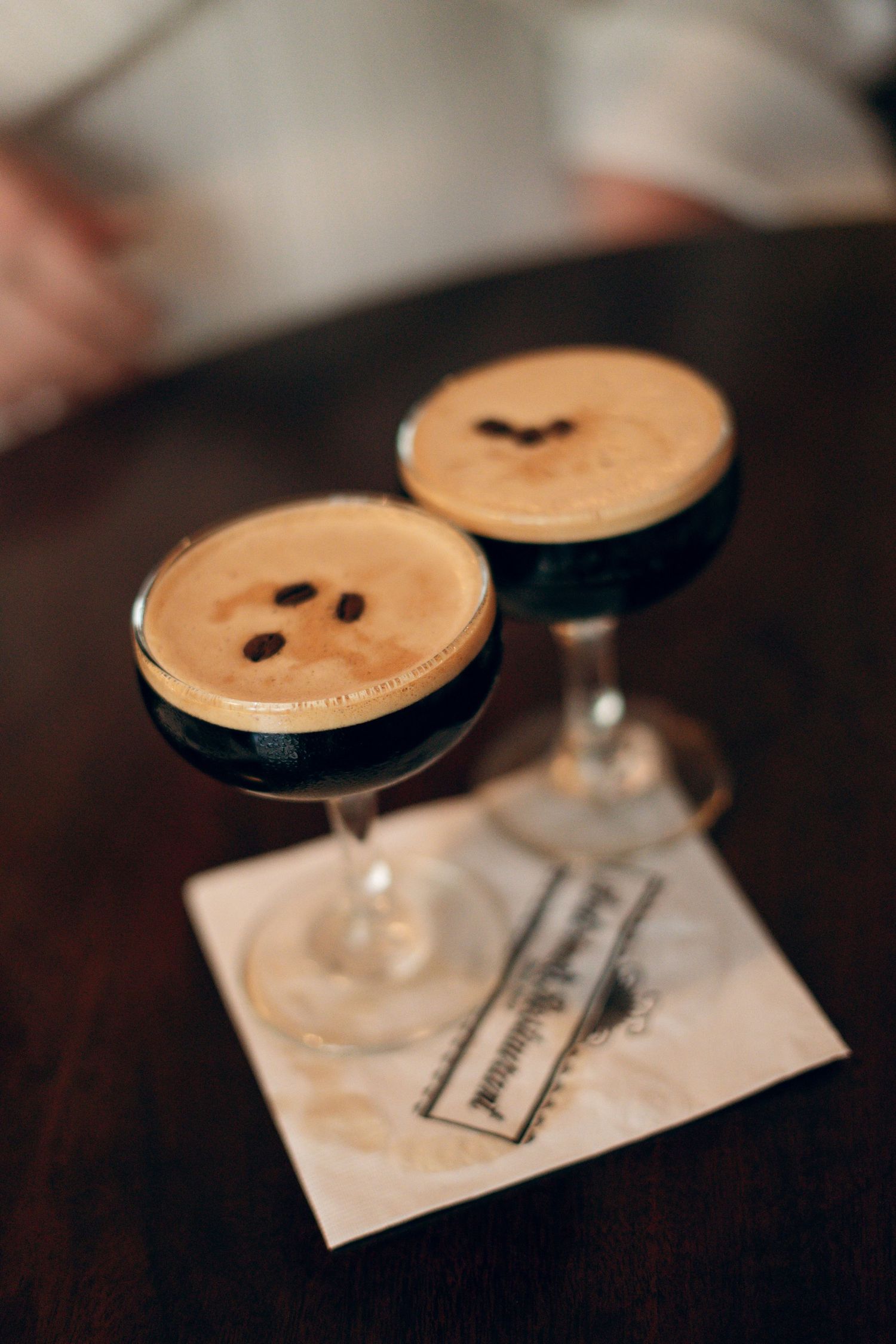 Two elegant espresso martinis with creamy foam tops and coffee beans garnish sit on napkin at dark wooden table.