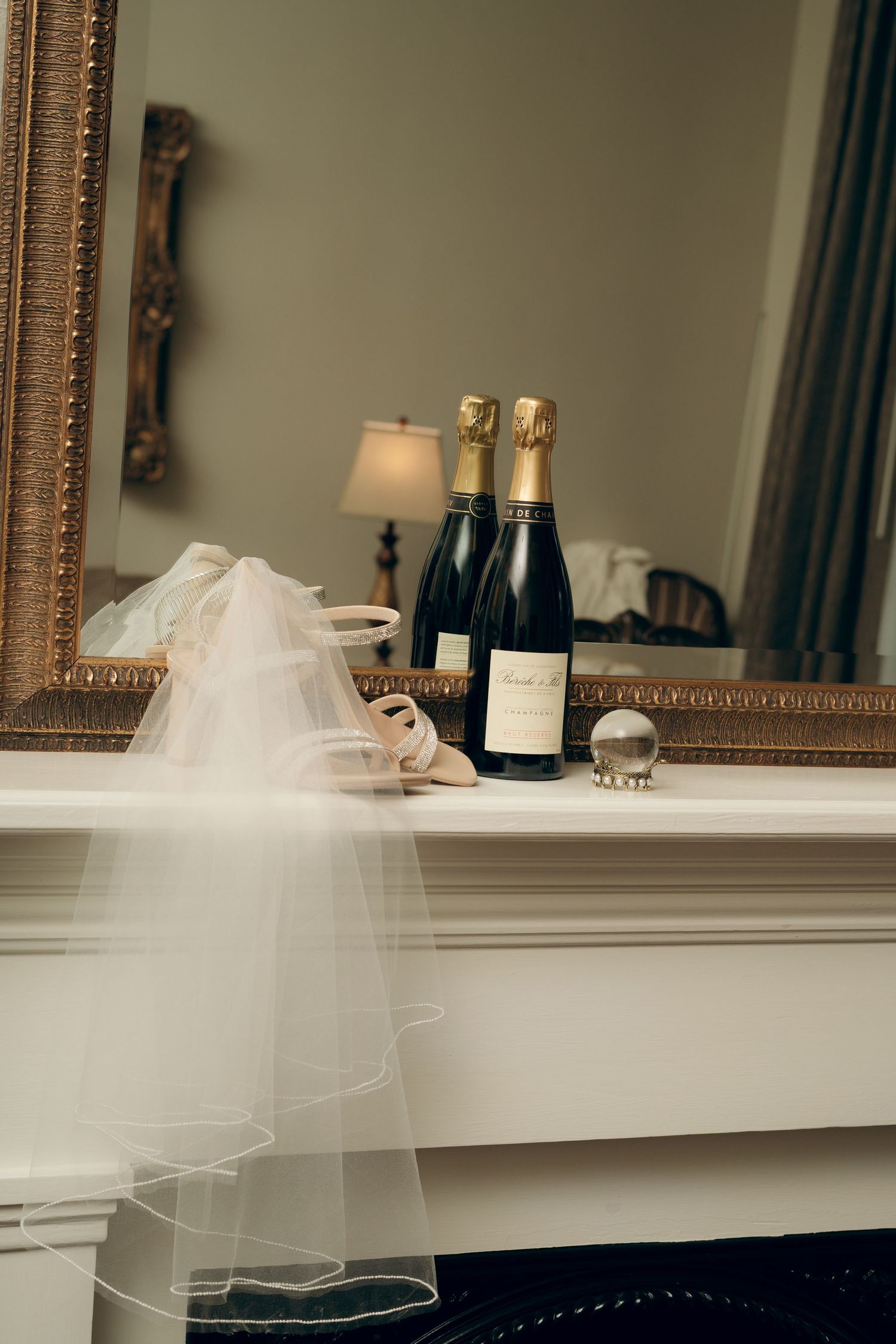 Elegant champagne bottles and bridal accessories displayed on a white mantel with ornate mirrors.