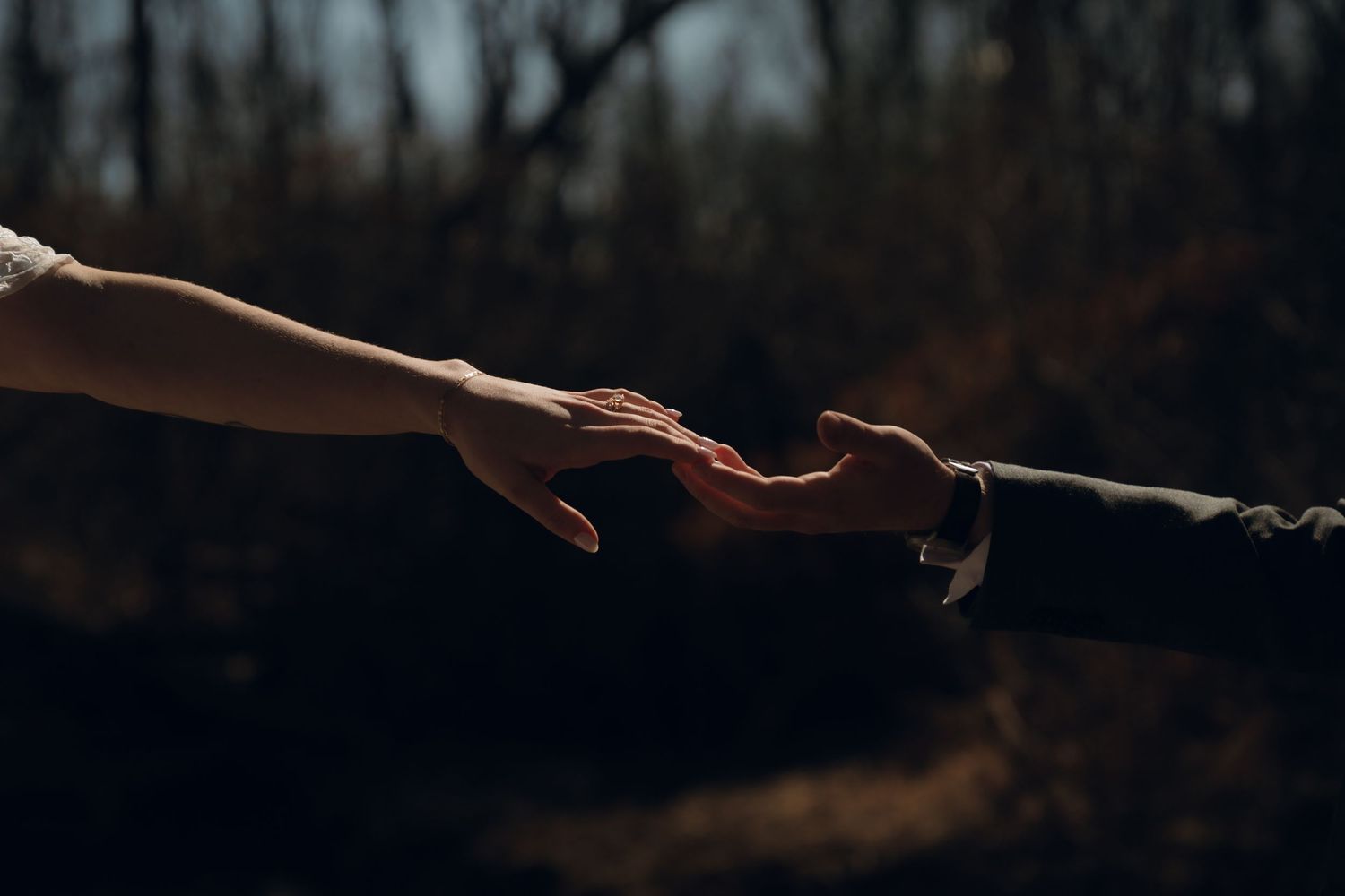 Two hands reaching toward each other in silhouette, creating an intimate connection against a dark blurred background.