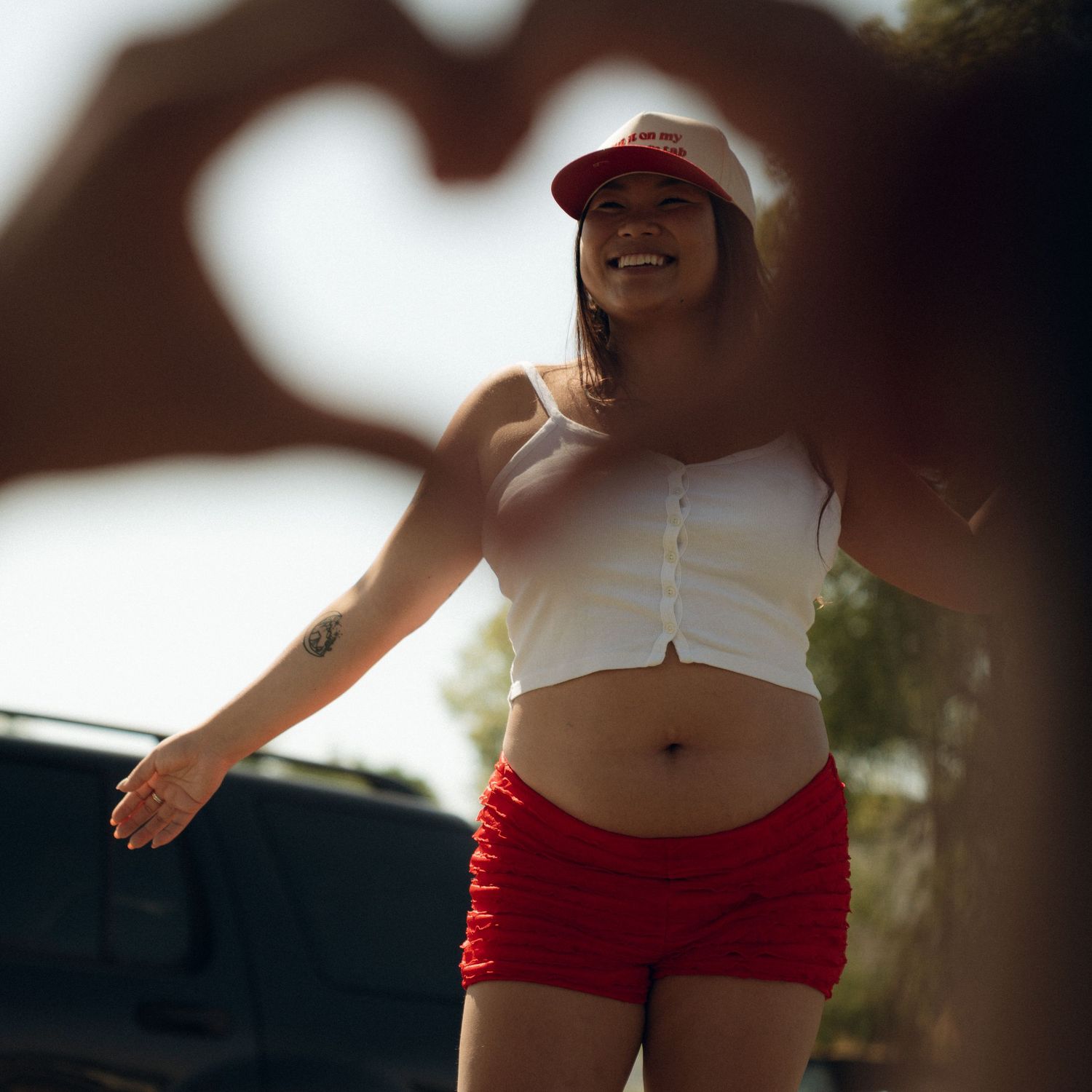 A person in red shorts and crop top stands by a car while hands in foreground form a heart shape silhouette.