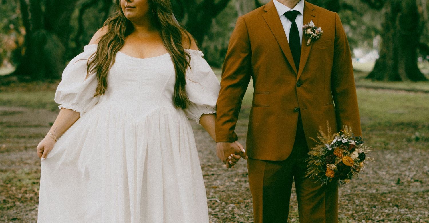 A couple walks together on a dirt path through autumn trees in formal attire.