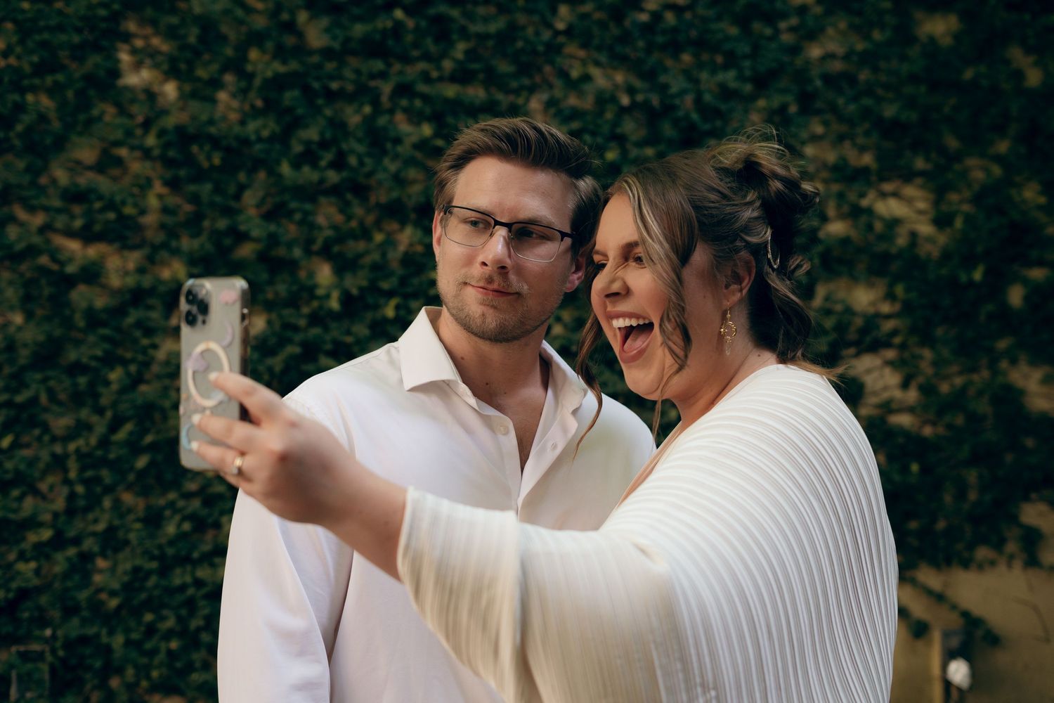 A joyful couple takes a selfie together outdoors with autumn foliage in the background.