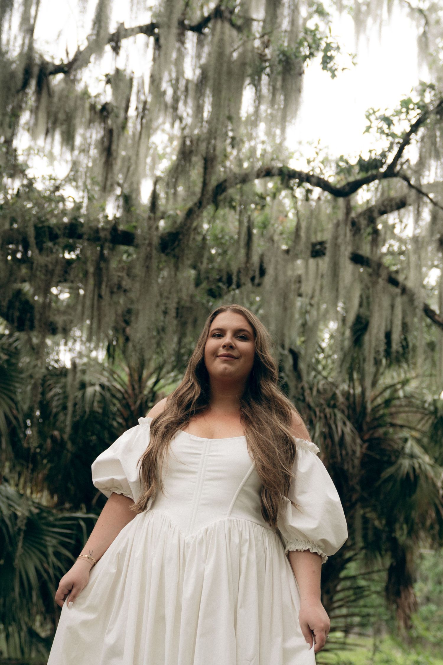 A dreamy bridal portrait shows a person in a white puff-sleeve dress surrounded by Spanish moss and palm trees.