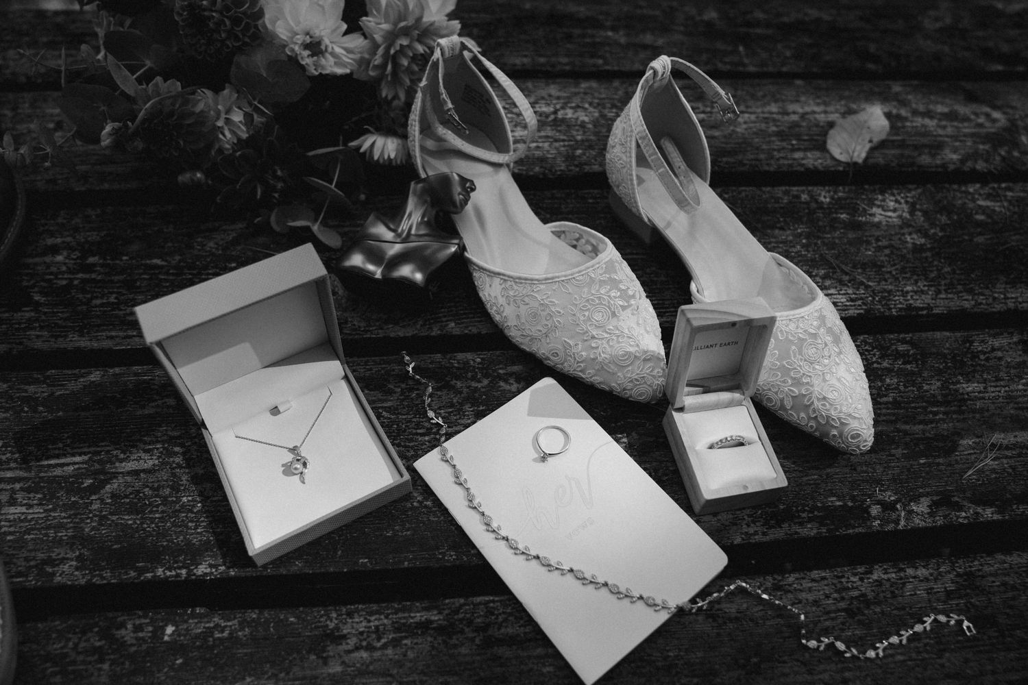 Elegant white lace bridal shoes with ankle straps displayed next to jewelry boxes on weathered wood surface.