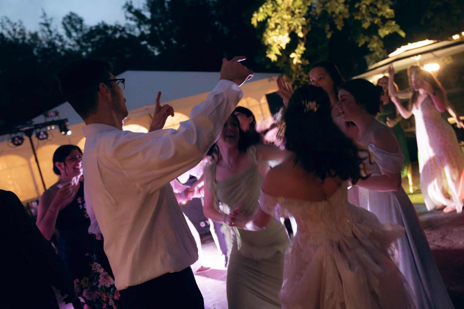 Party guests dance energetically at a nighttime outdoor wedding reception with pink and purple lighting effects.