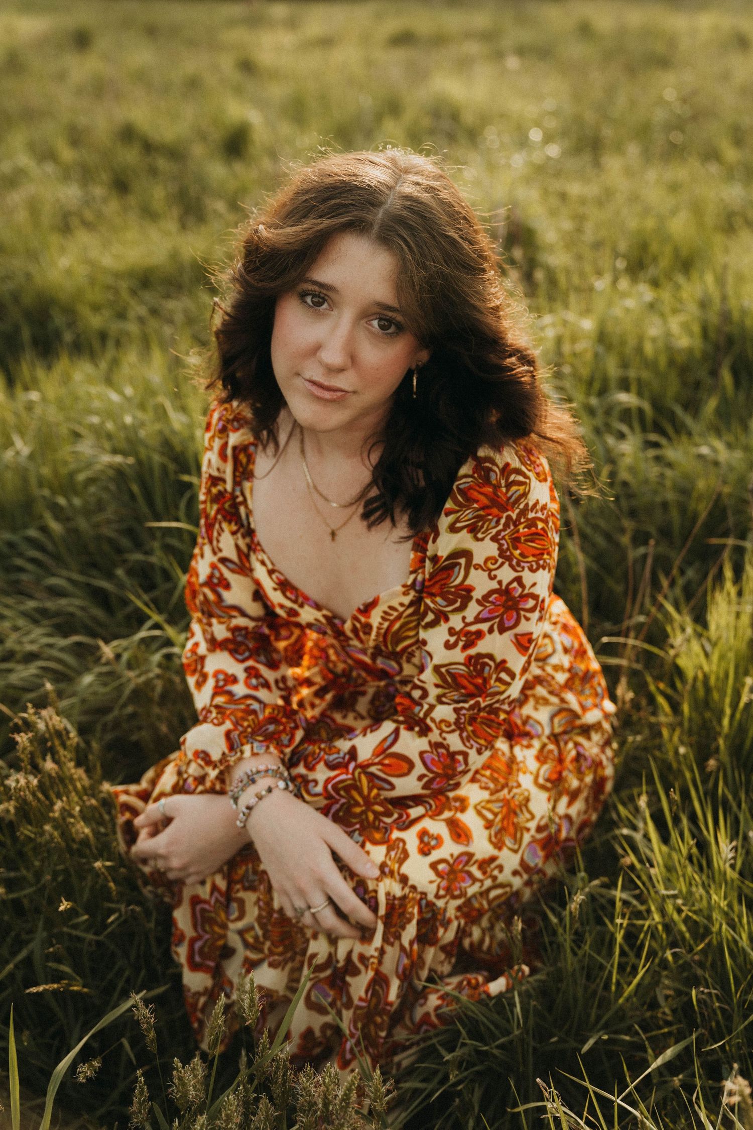 Woman in vintage-style floral orange dress poses in golden grassy field at sunset.