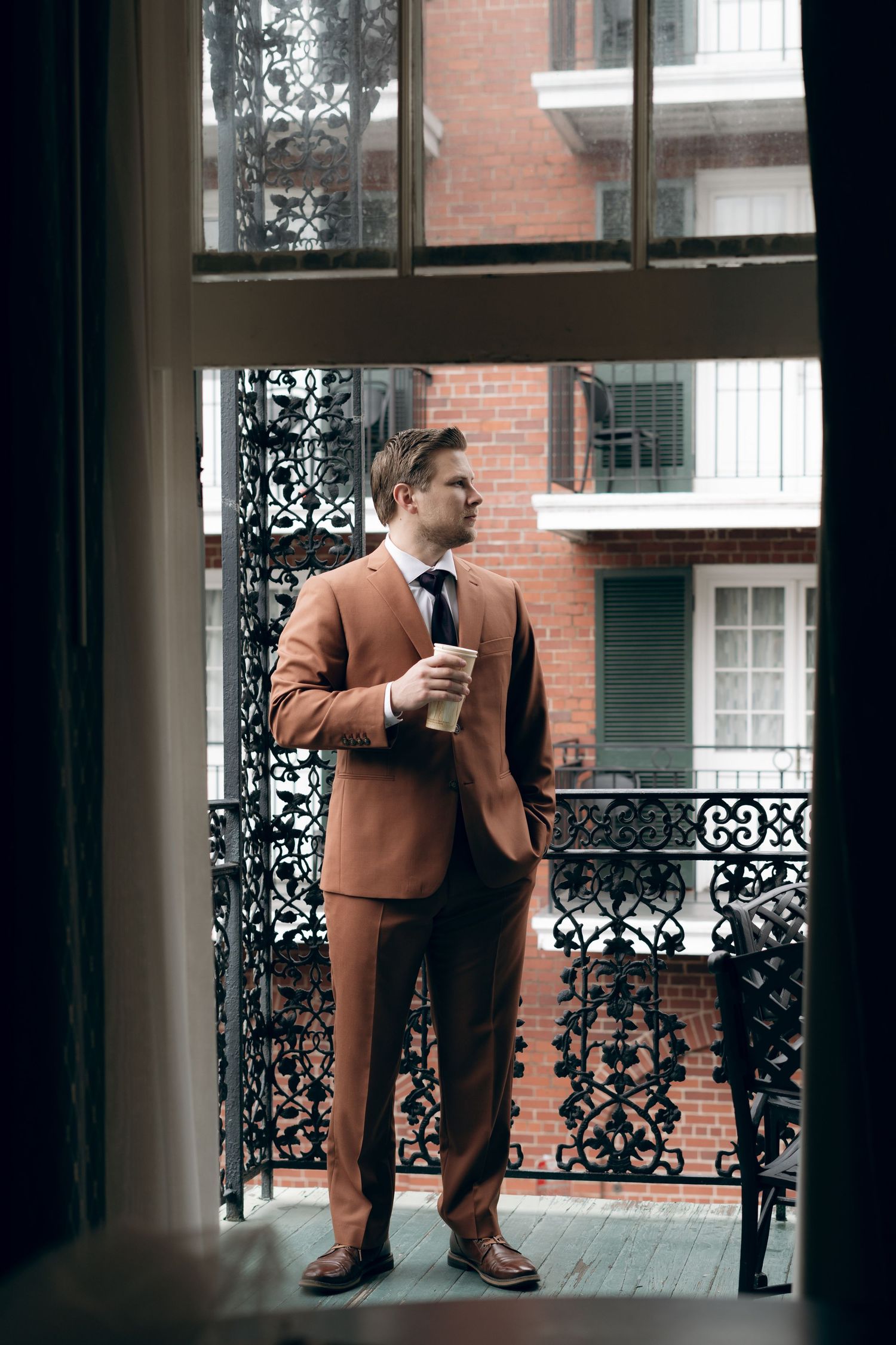 A person in a brown suit holds a coffee cup while standing on an ornate balcony overlooking brick buildings.