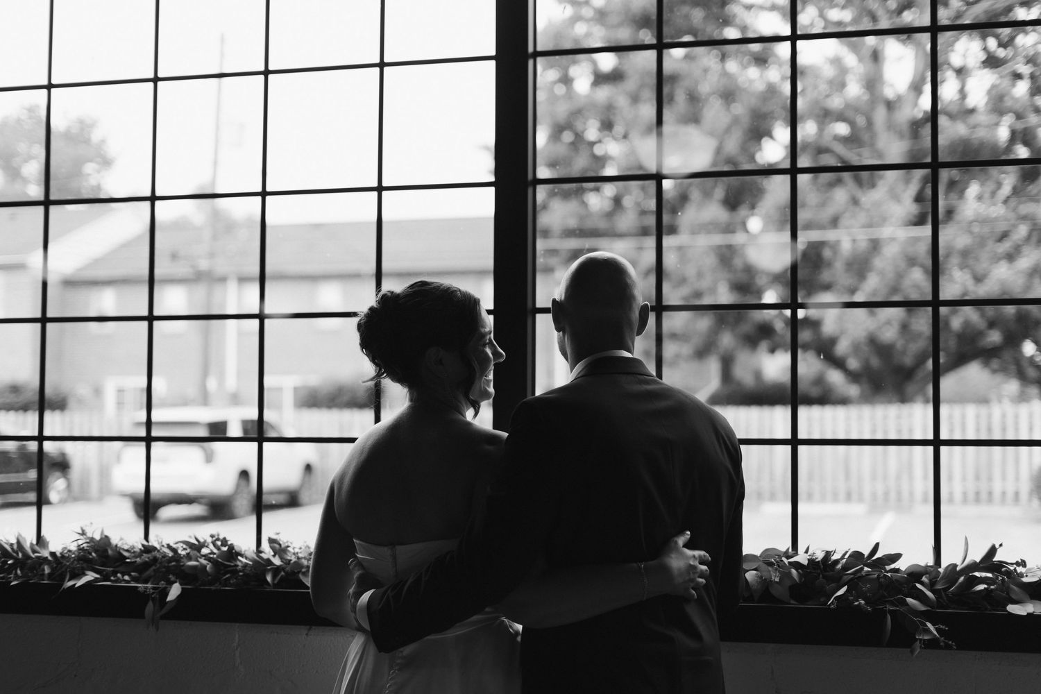 Black and white photo of a couple embracing while looking out a large window at their wedding venue.