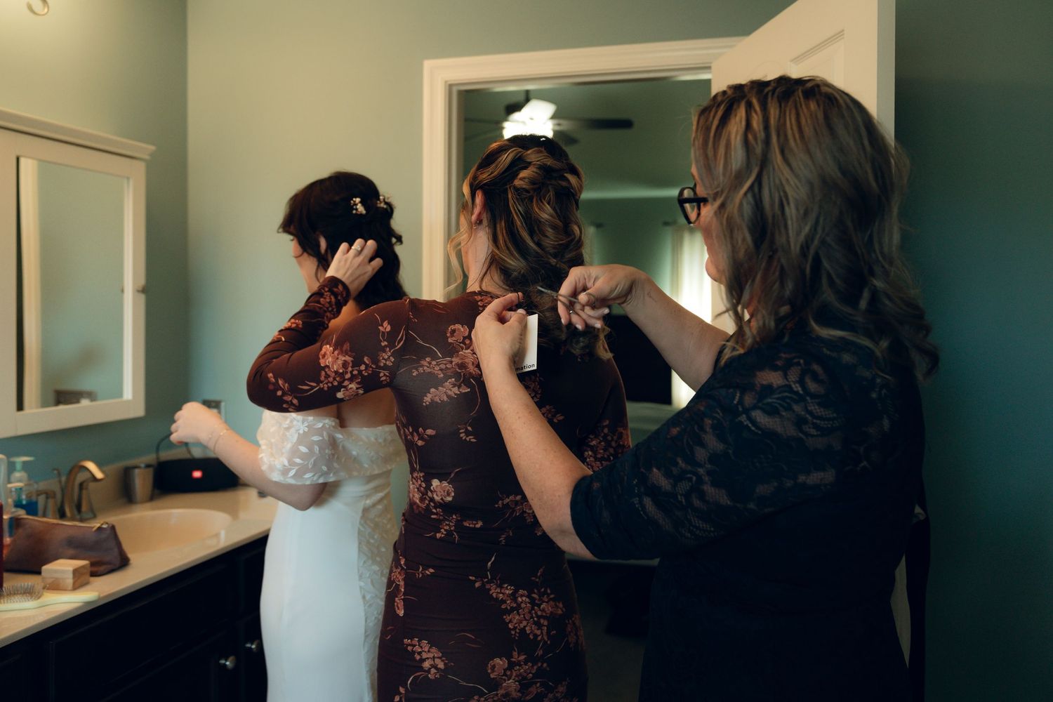 Two women in a bathroom help adjust a wedding dress during bridal preparations.