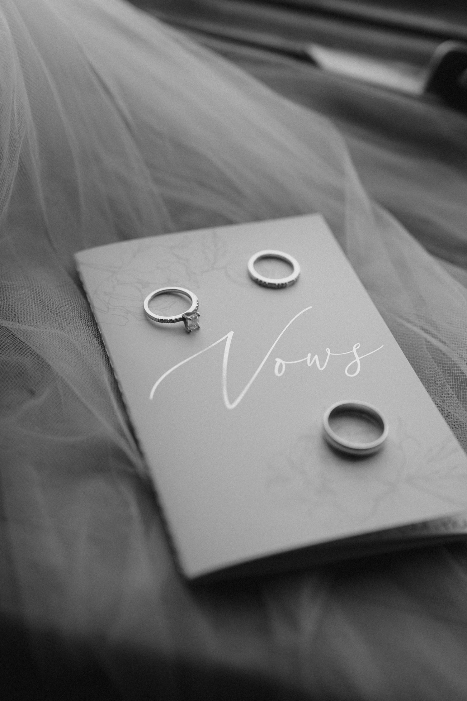 Wedding rings and vows card arranged on flowing tulle fabric in elegant black and white photography.