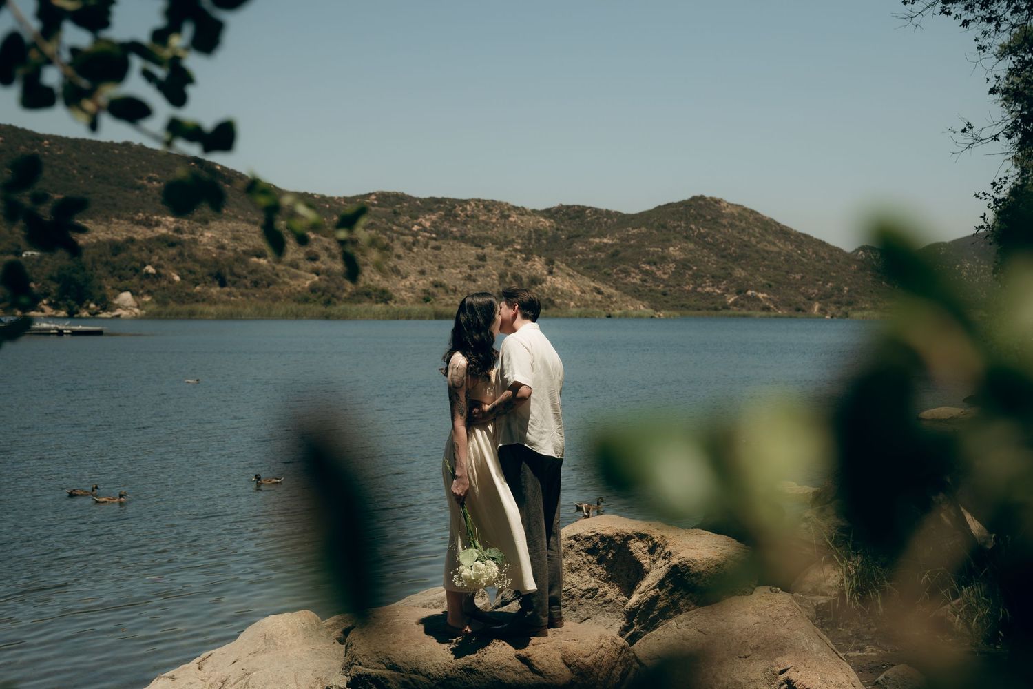 Romantic couple embraces on rocky shoreline overlooking serene mountain lake during golden afternoon light.