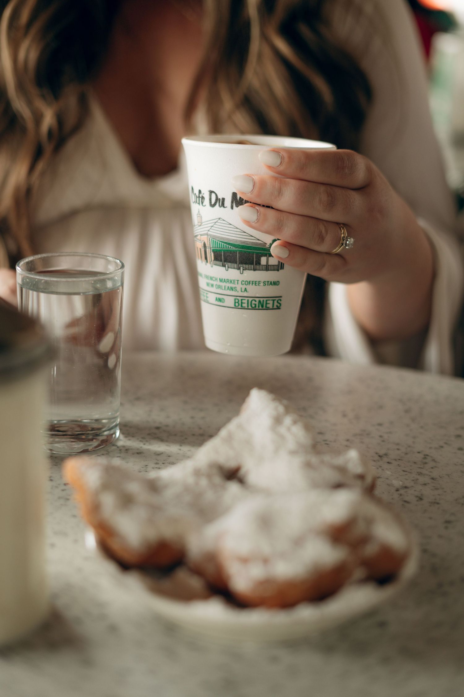 A hand holds a takeout coffee cup next to a plate of powdered beignets and a glass of water on a marble counter.