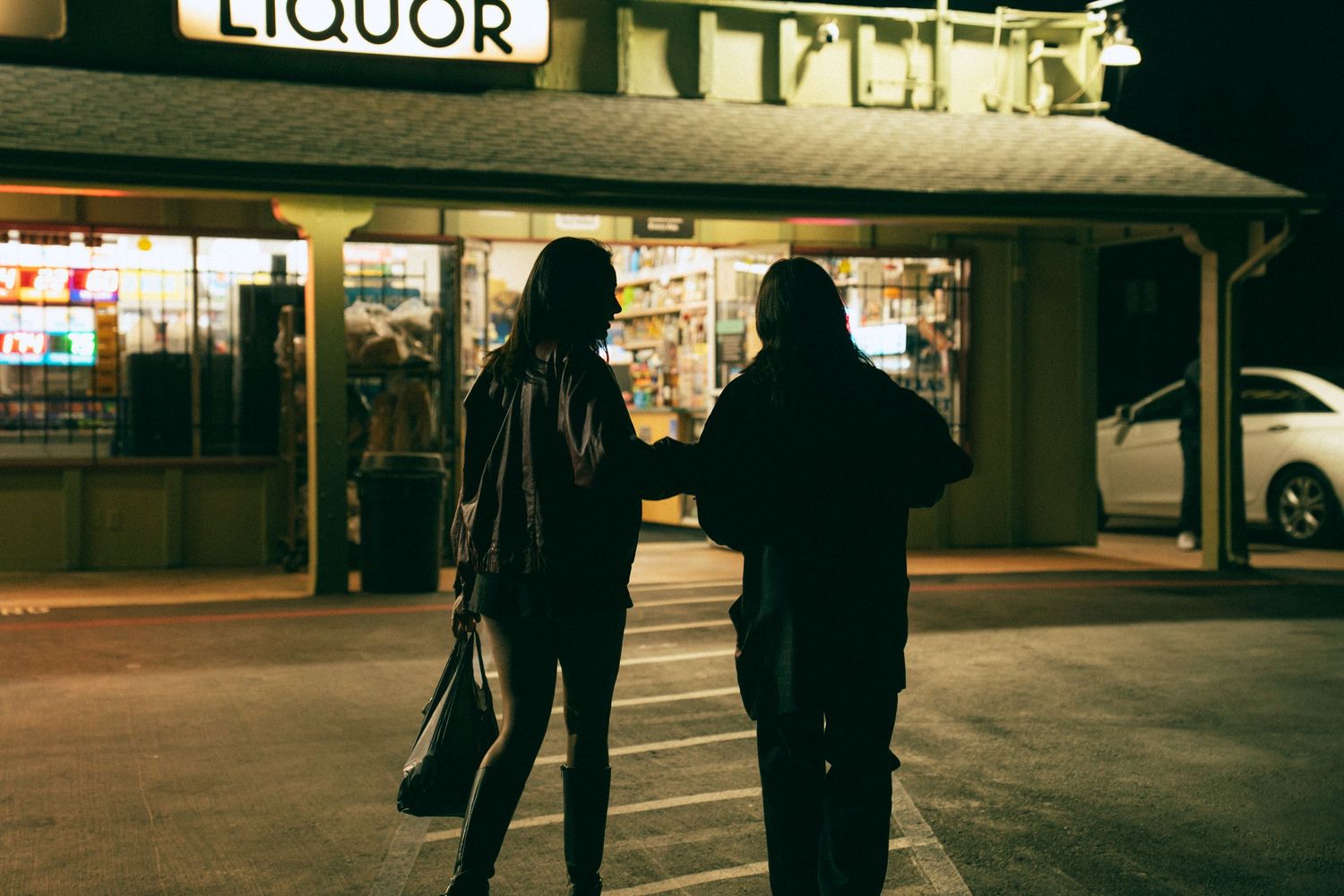Dark silhouettes stand beneath a liquor store sign in a moody nighttime street scene.