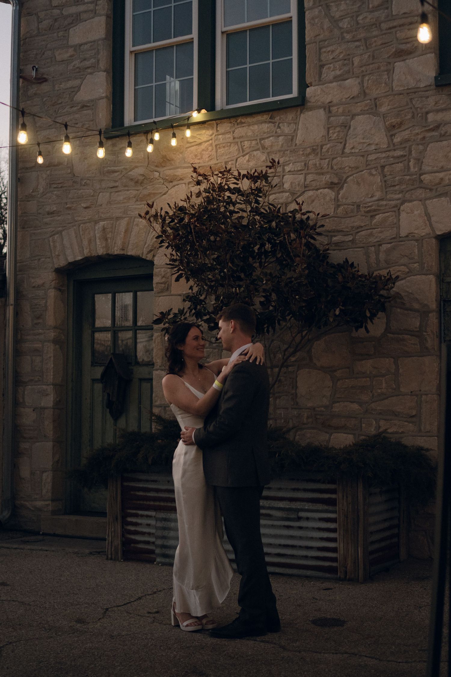 A romantic couple embraces outside a stone building with string lights glowing above at dusk.
