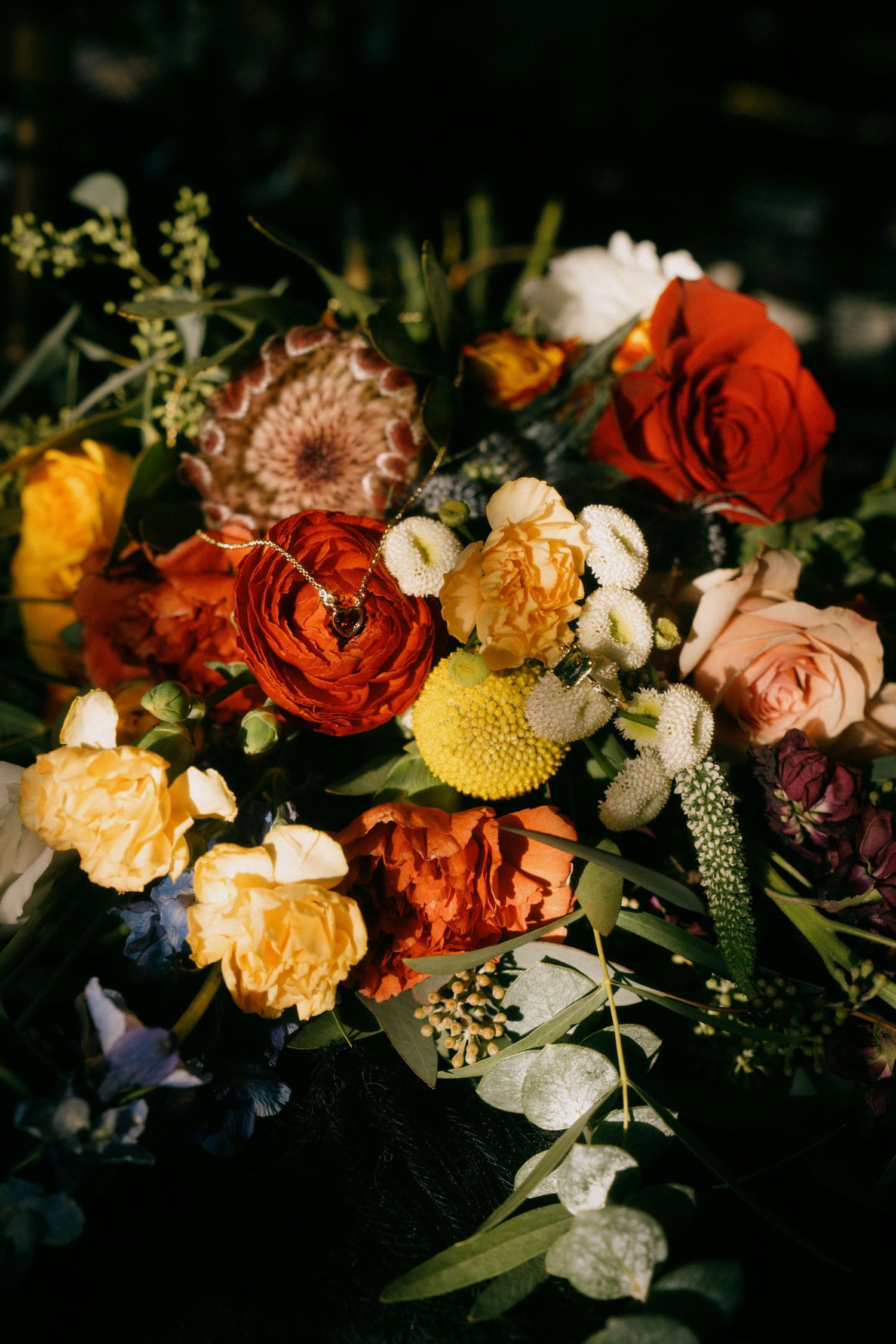 A romantic floral arrangement featuring deep red roses, peach blooms, and yellow flowers with moody lighting and dark shadows.