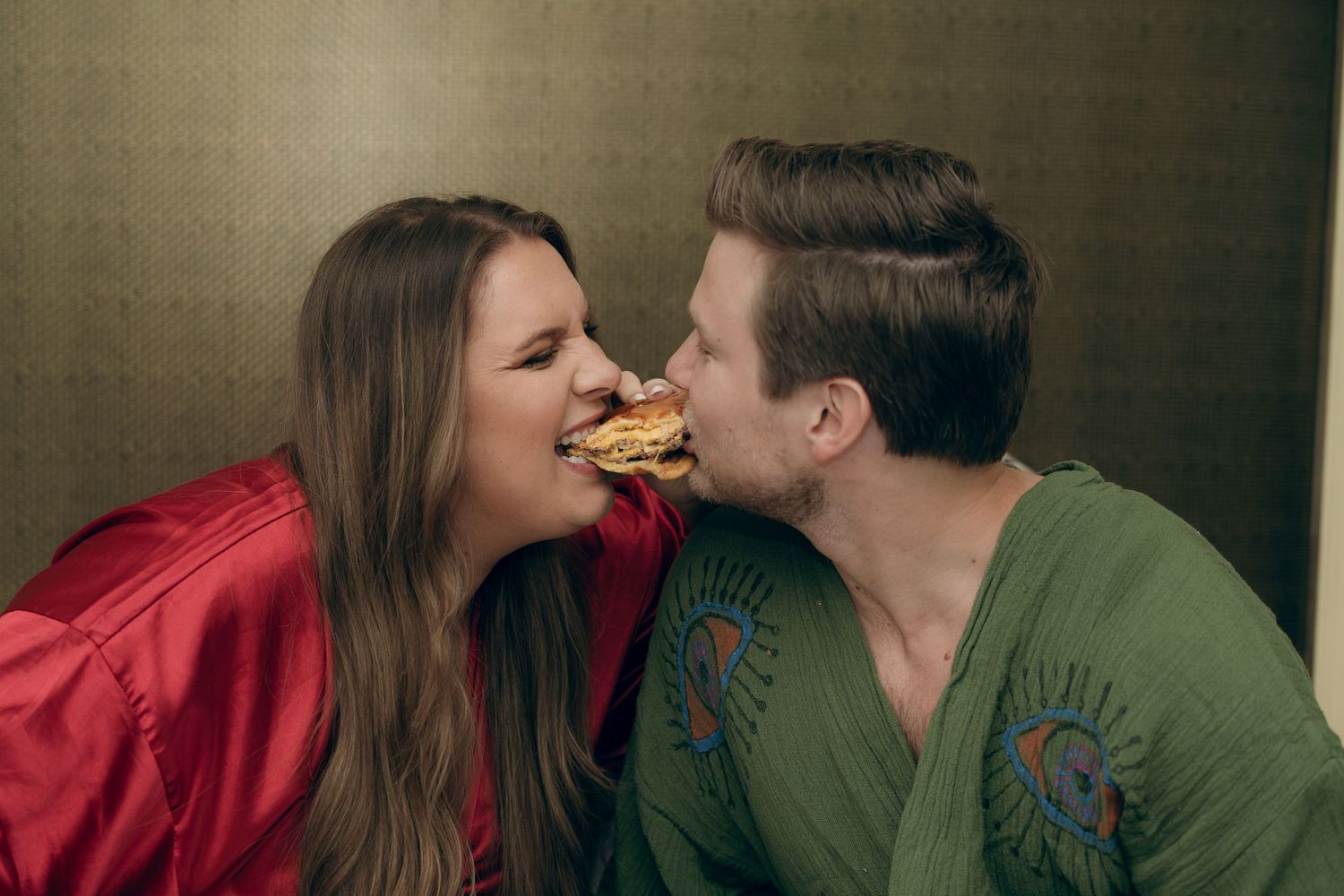 Two people playfully sharing a hamburger while wearing red and green tops in a casual indoor setting.