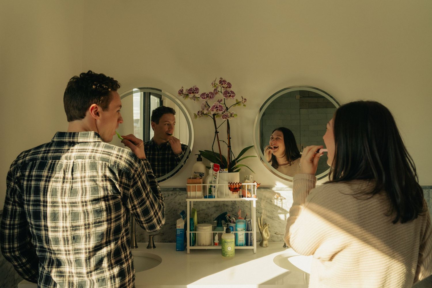 People gather around a mirror and bathroom counter in casual home setting.