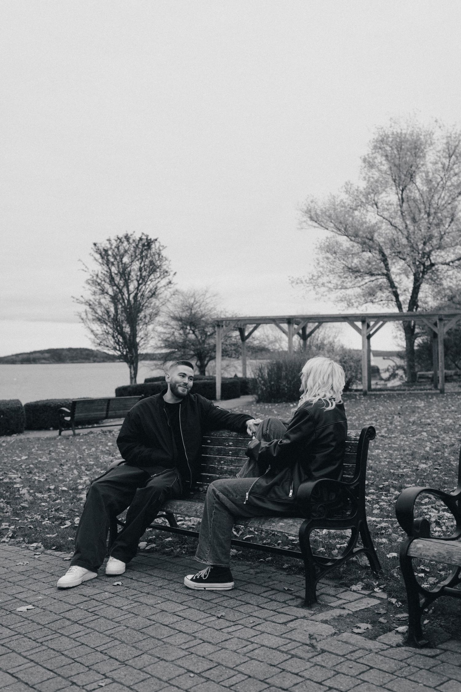 Two people sit conversing on a park bench with bare trees and waterfront views in the background. Couple in Bar Harbor Maine